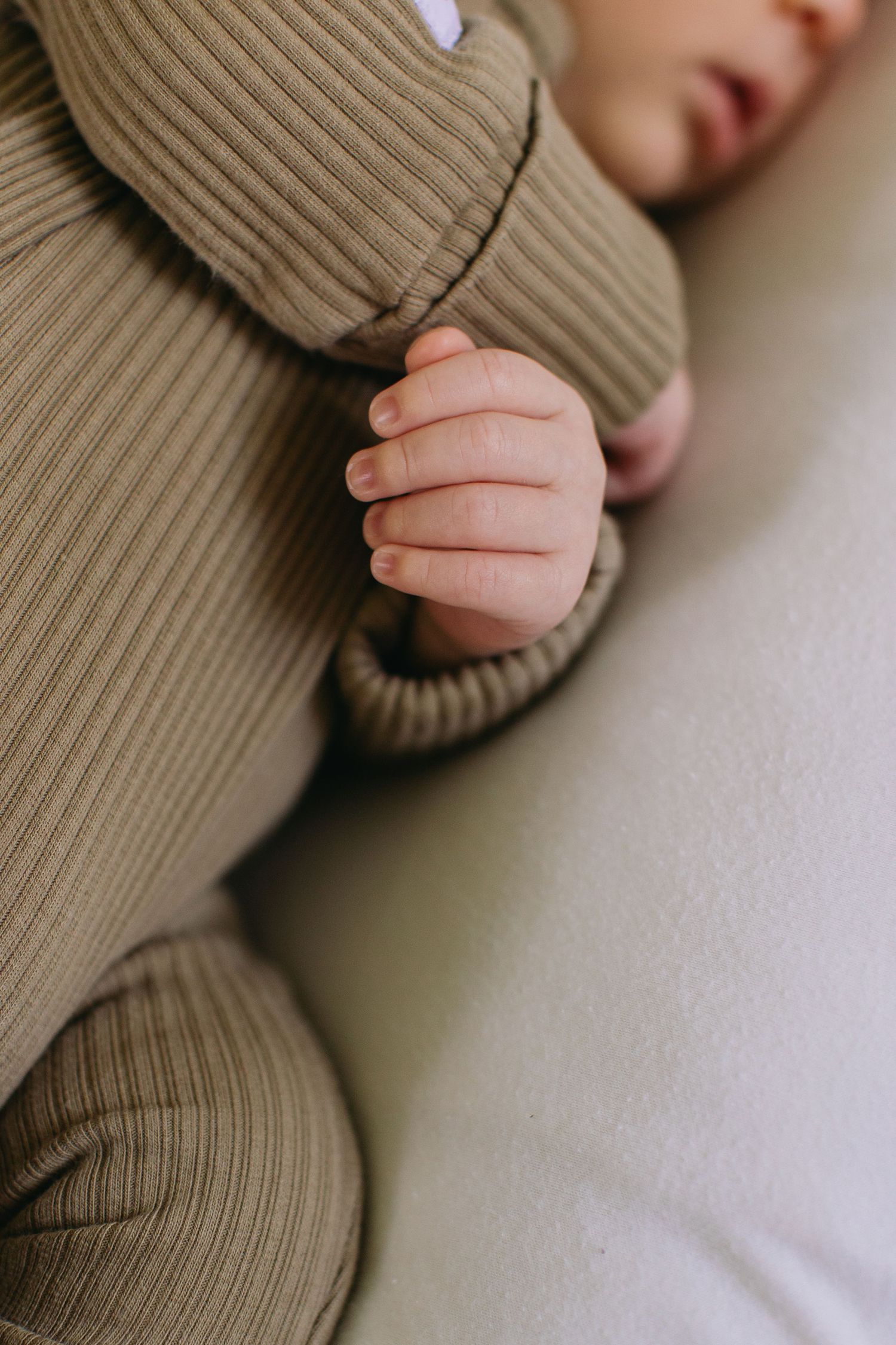 Close-up of a sleeping baby's tiny hand in a ribbed olive green romper on a soft white surface.