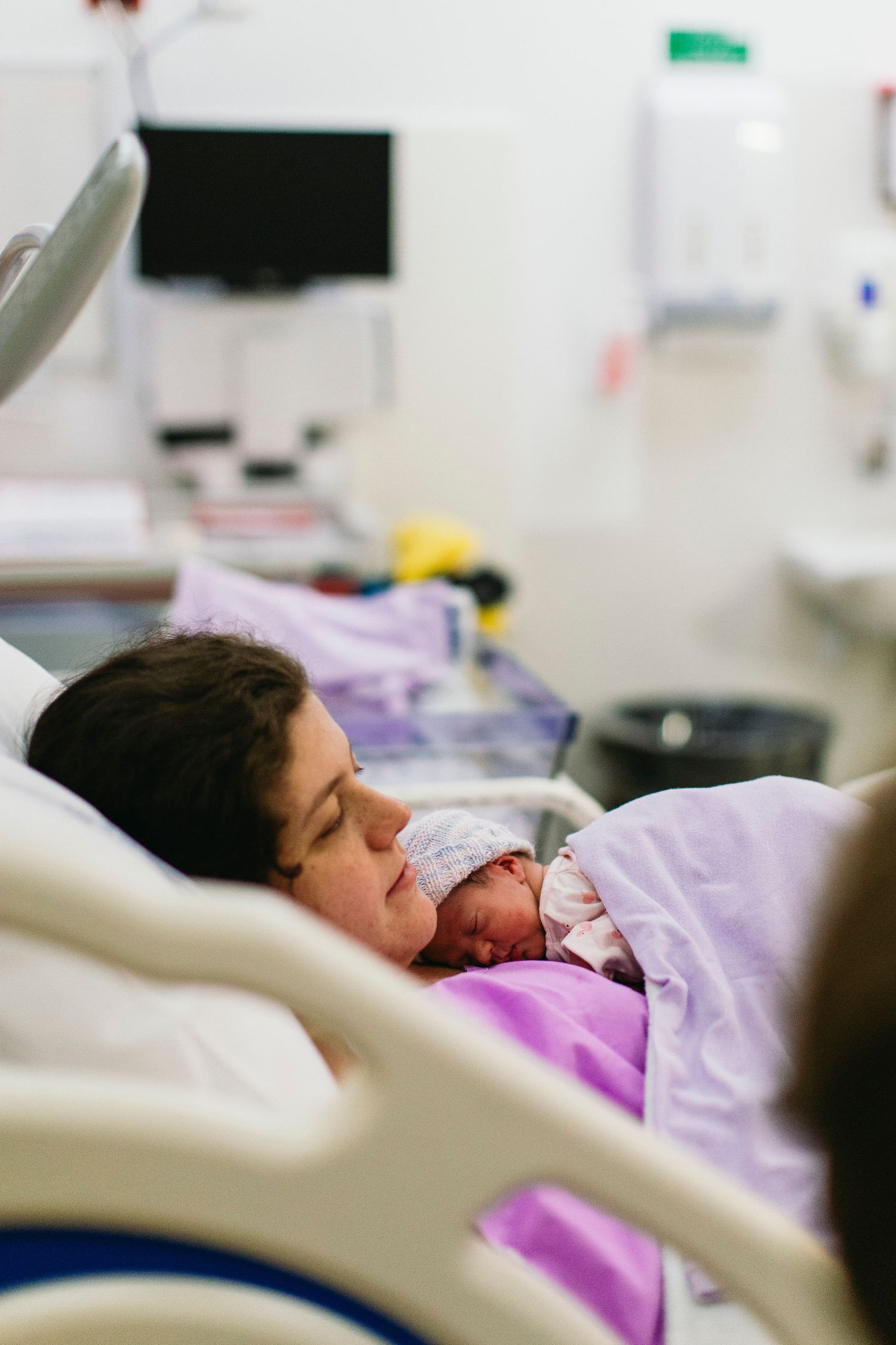 New mother lying in hospital bed gazing lovingly at her newborn baby wrapped in a white blanket.