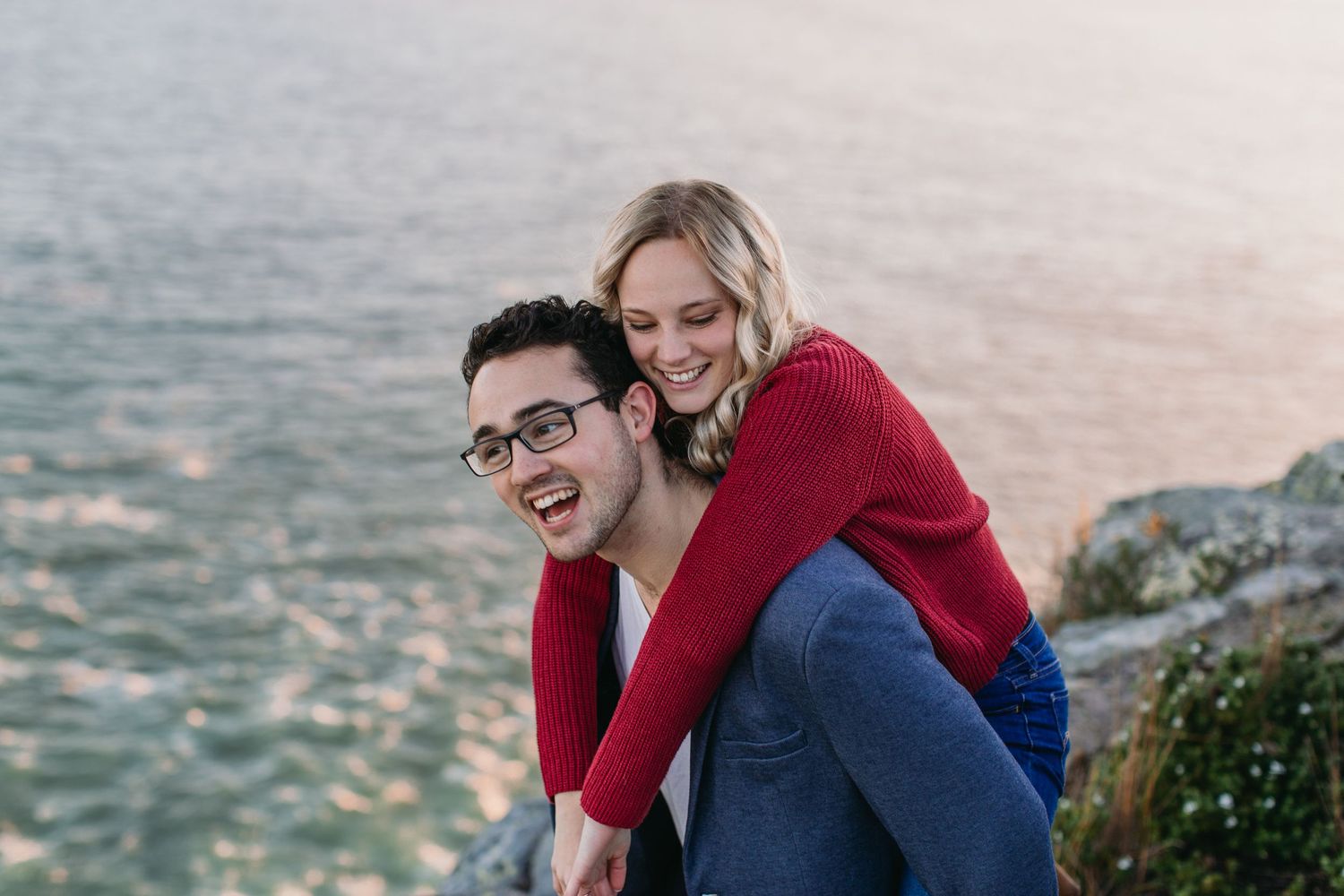 A smiling couple poses outdoors near the ocean, woman in red top giving a piggyback ride to a man wearing glasses.