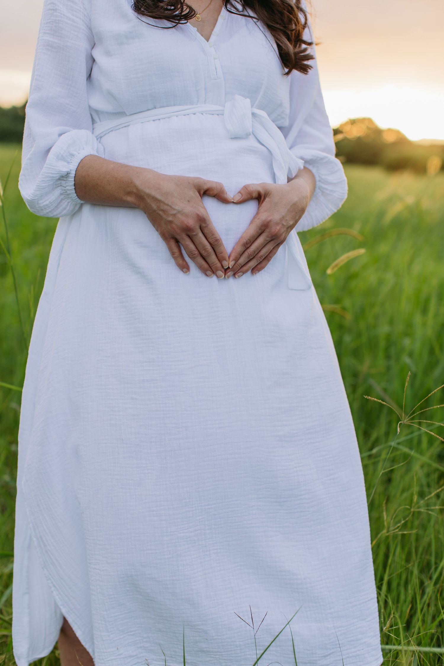 Pregnant woman in white dress forming heart shape with hands on baby bump in green field at sunset.