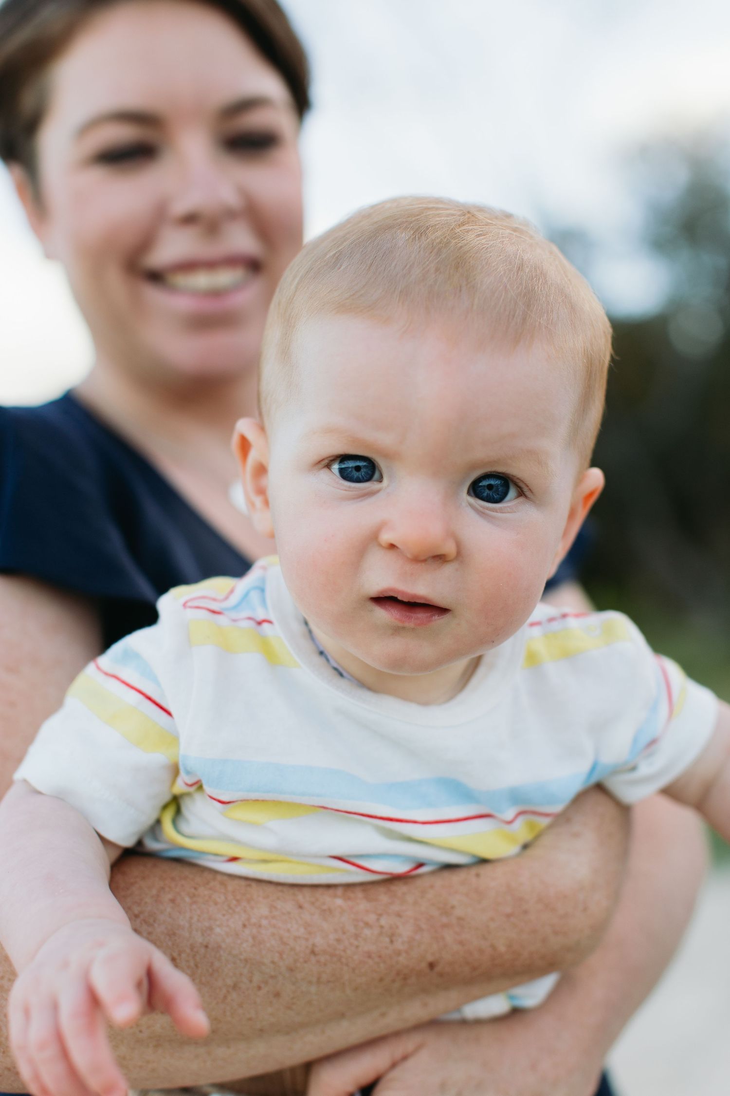A smiling woman holds a curious blue-eyed baby in a striped onesie outdoors.