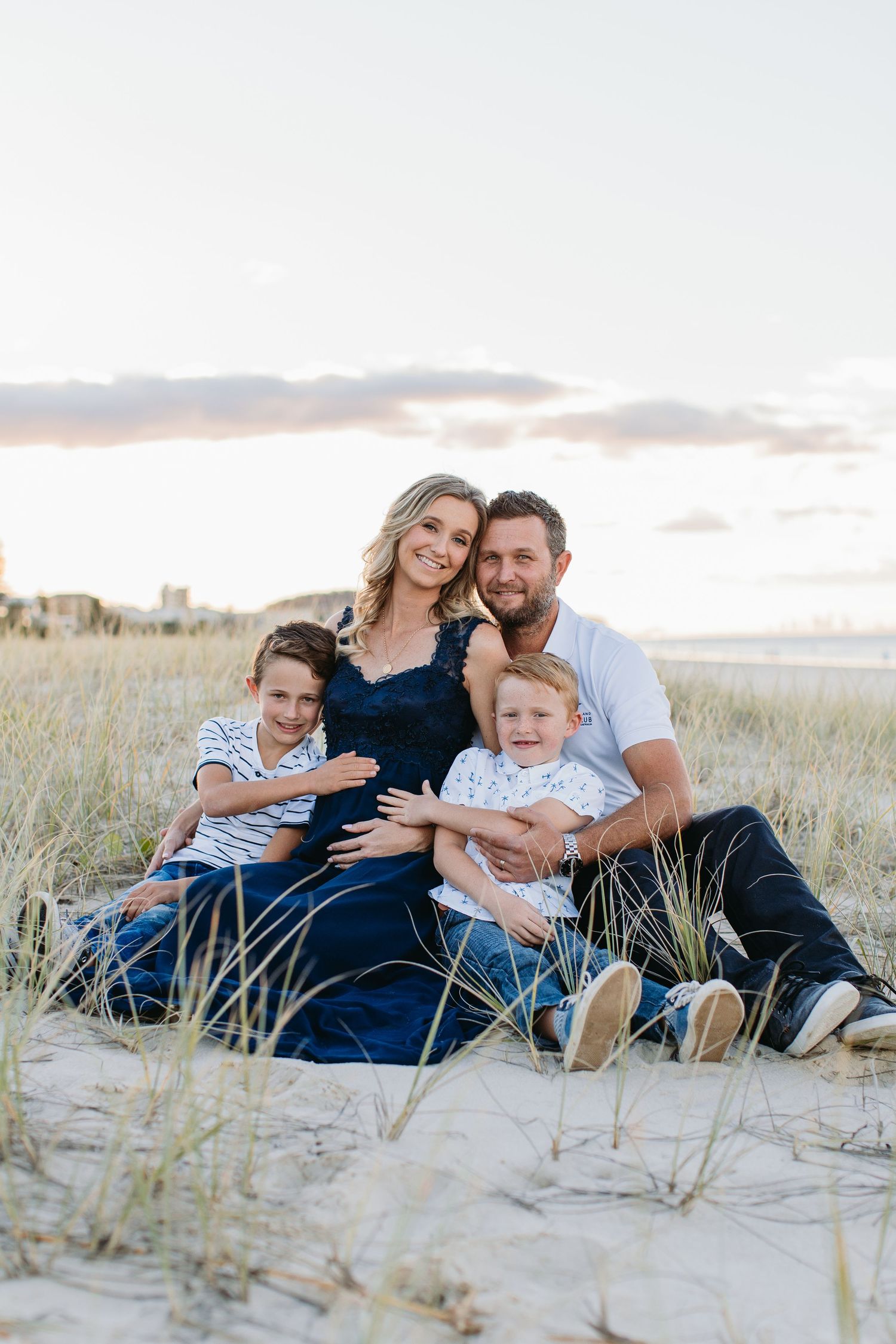 Family of four poses on sandy beach dunes, mother in navy dress, father in white shirt, two young boys smiling.