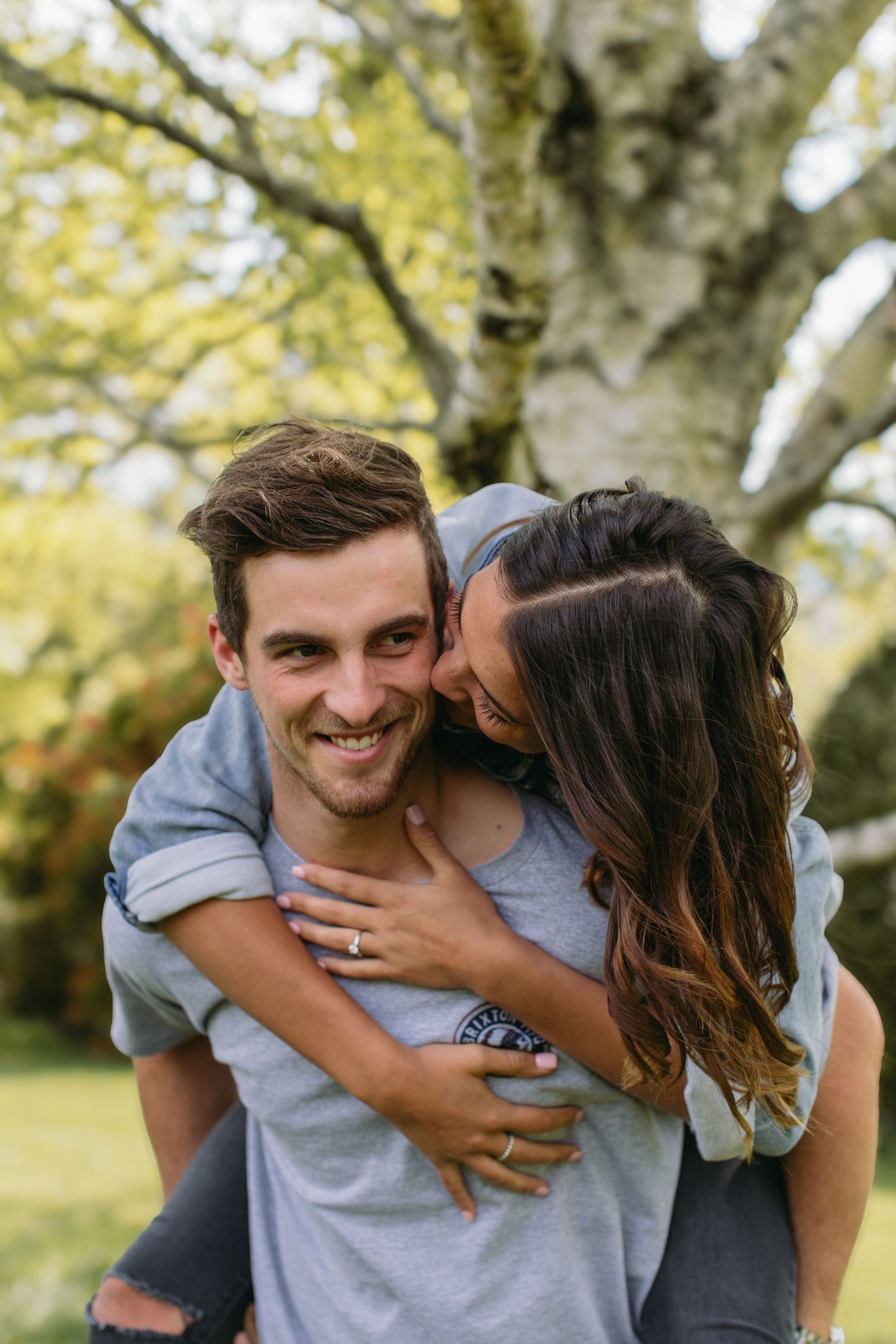 A smiling man gives a woman a piggyback ride outdoors, as she kisses his cheek wearing an engagement ring.
