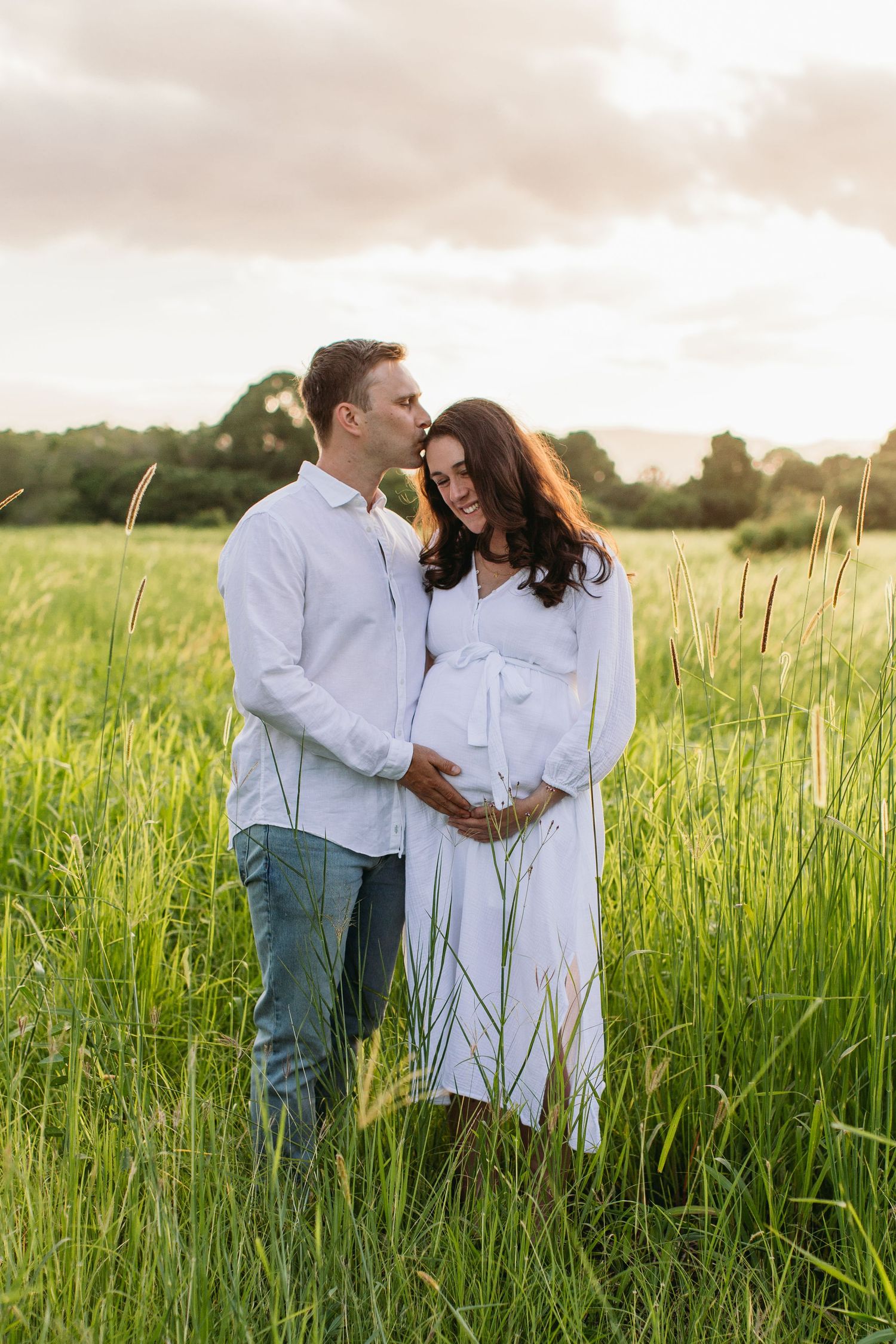 A couple stands in a lush green field at sunset, man kisses pregnant woman's head as she cradles her baby bump.