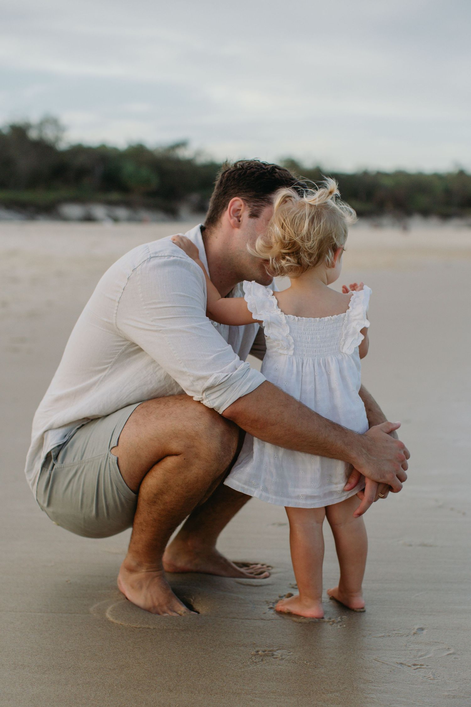 Father crouching beside toddler daughter on sandy beach, both wearing white, sharing a tender moment by the ocean.