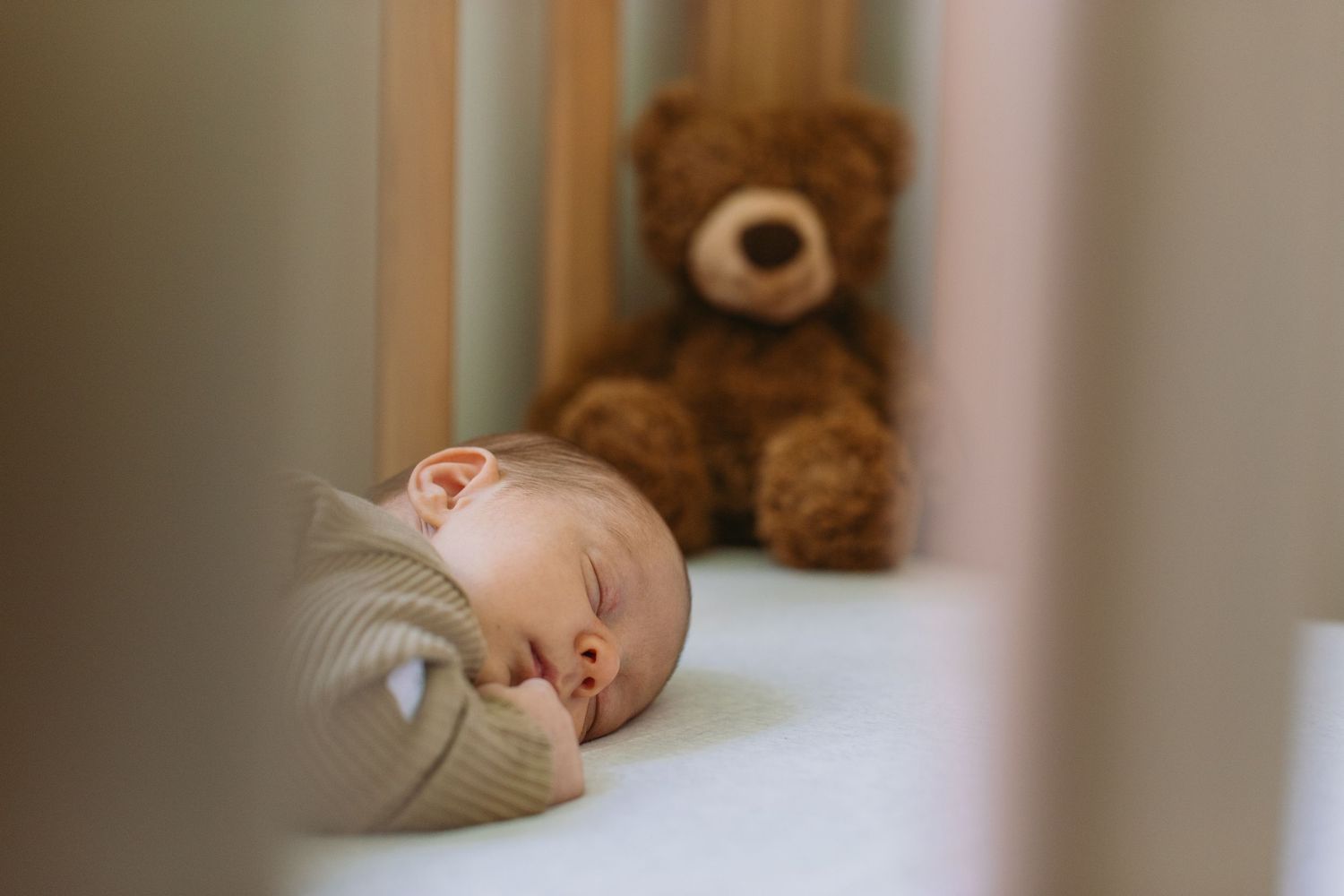 Newborn baby sleeping peacefully in a white crib beside a soft brown teddy bear.