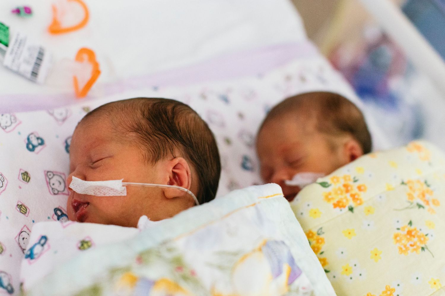 Two premature newborn twins sleeping side by side in NICU with oxygen tubes, wrapped in colorful blankets.