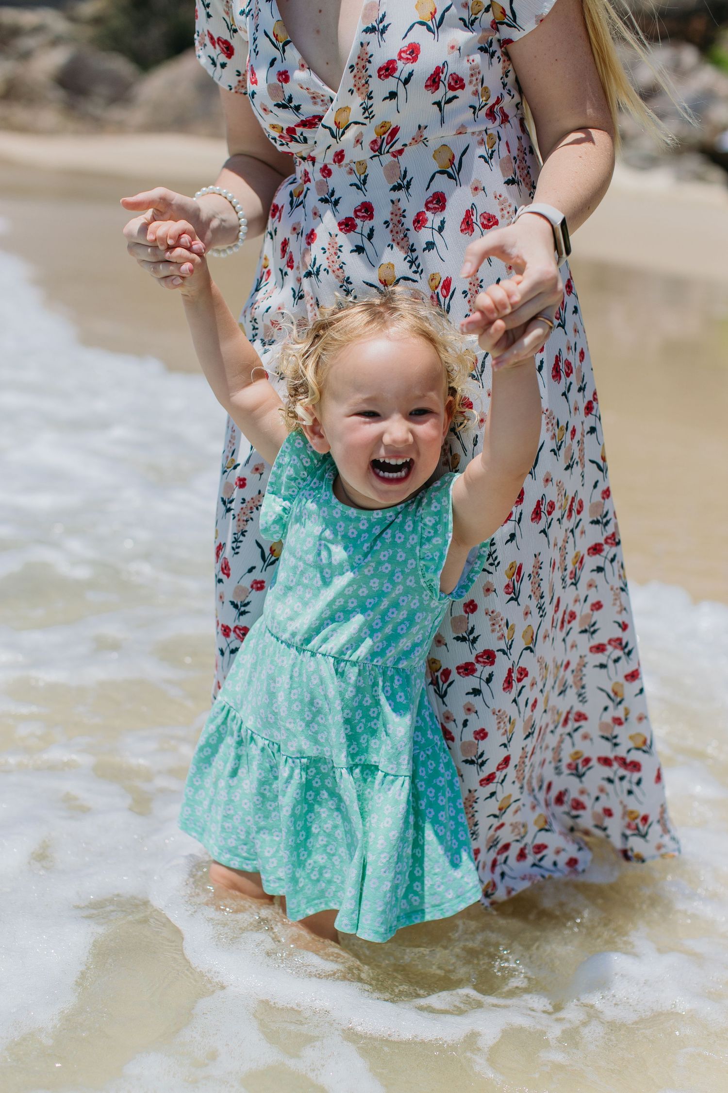 Excited toddler girl in teal dress laughing while mother holds her hands in ocean waves.
