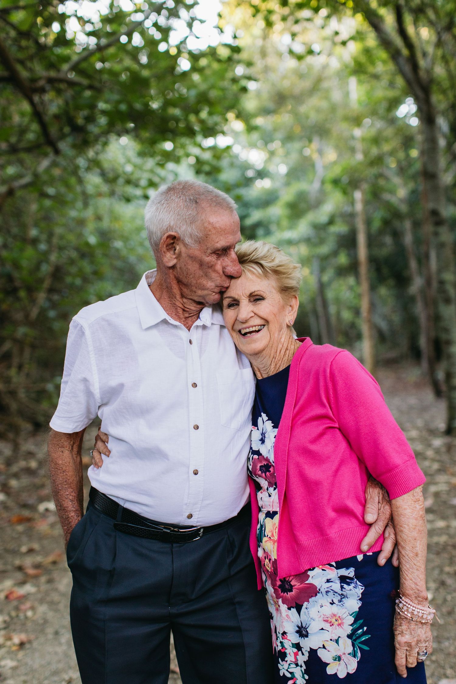 Elderly couple sharing a tender kiss outdoors, woman in pink cardigan laughing joyfully amid lush green trees.