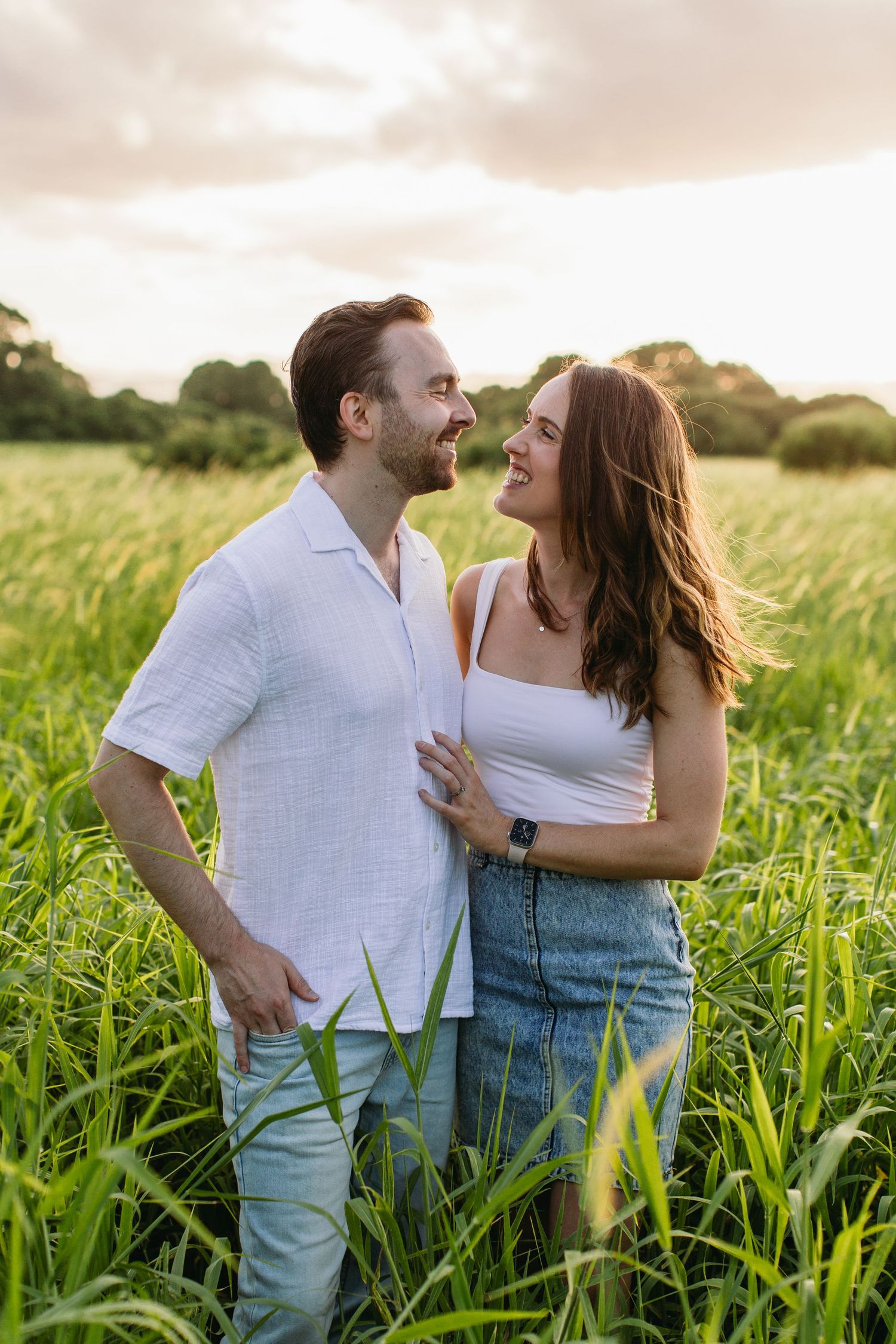A couple smiles at each other while standing in a lush green field at golden hour, man in white shirt and woman in denim skirt.