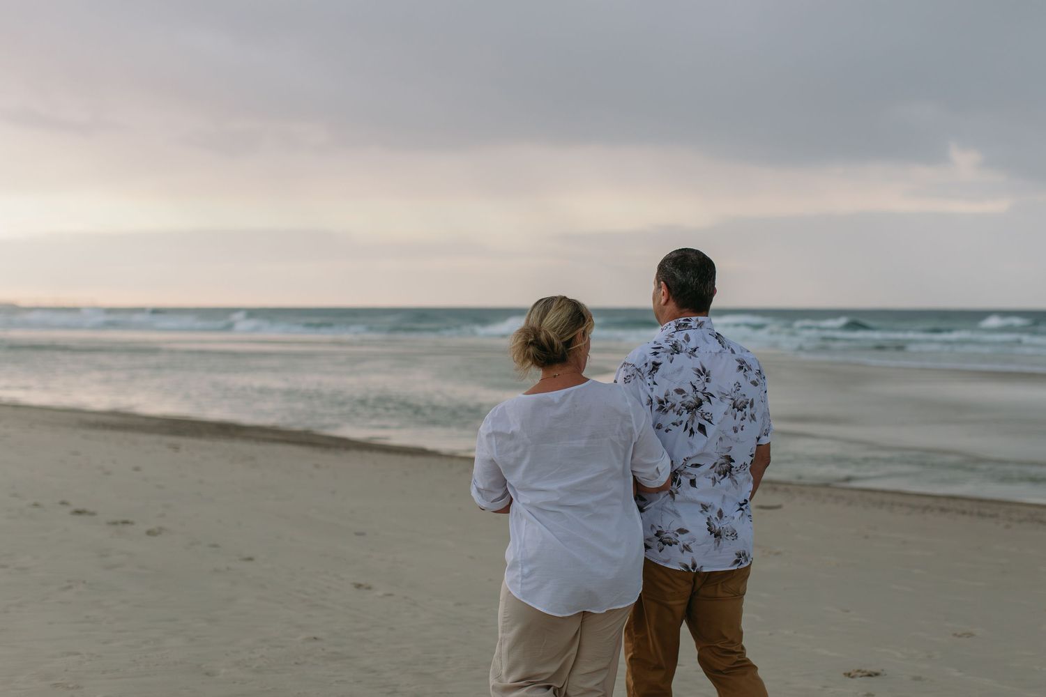 Elderly couple standing together on a sandy beach, gazing at the ocean waves under a cloudy sky.