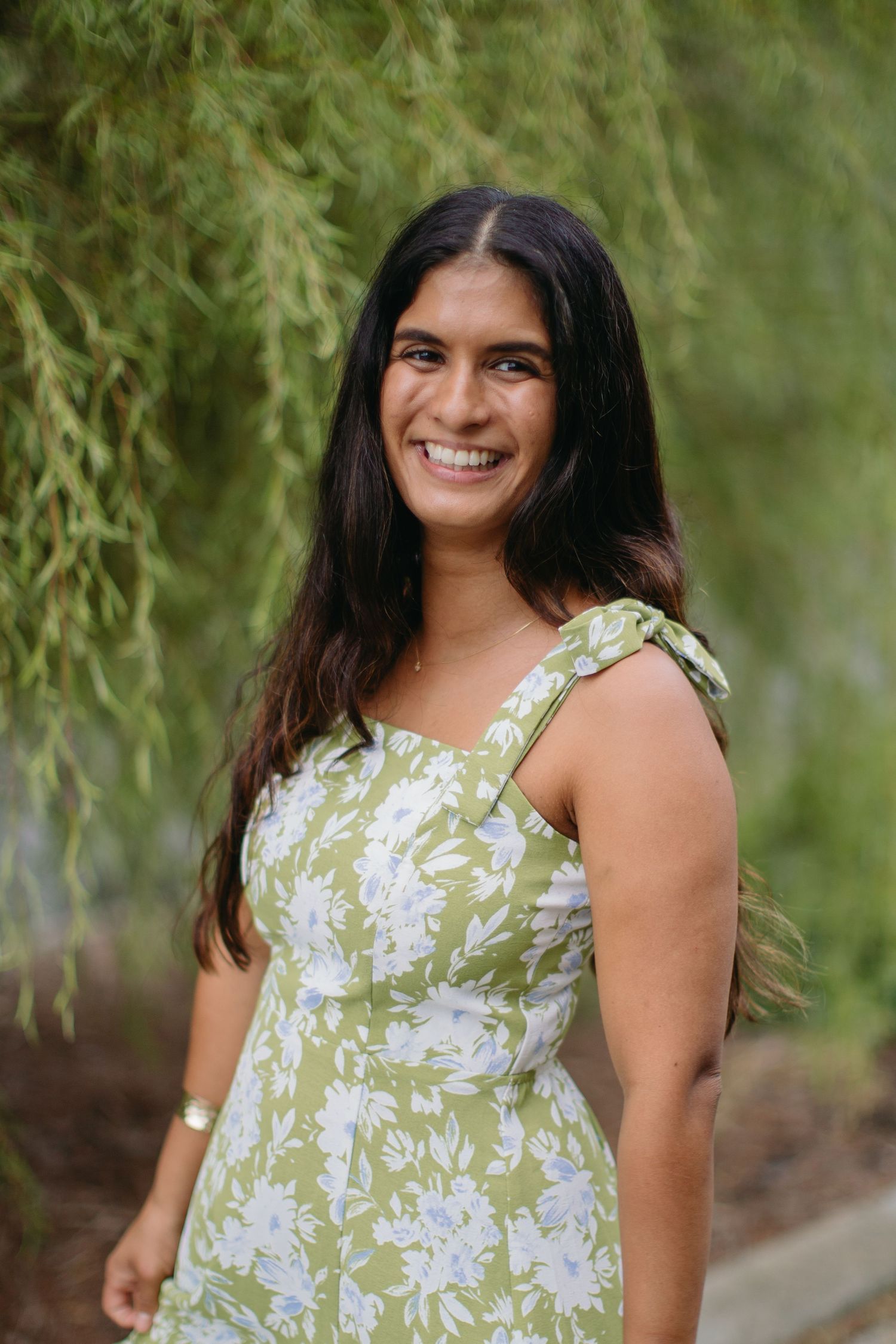 Young woman in green floral sundress smiling outdoors under weeping willow tree.