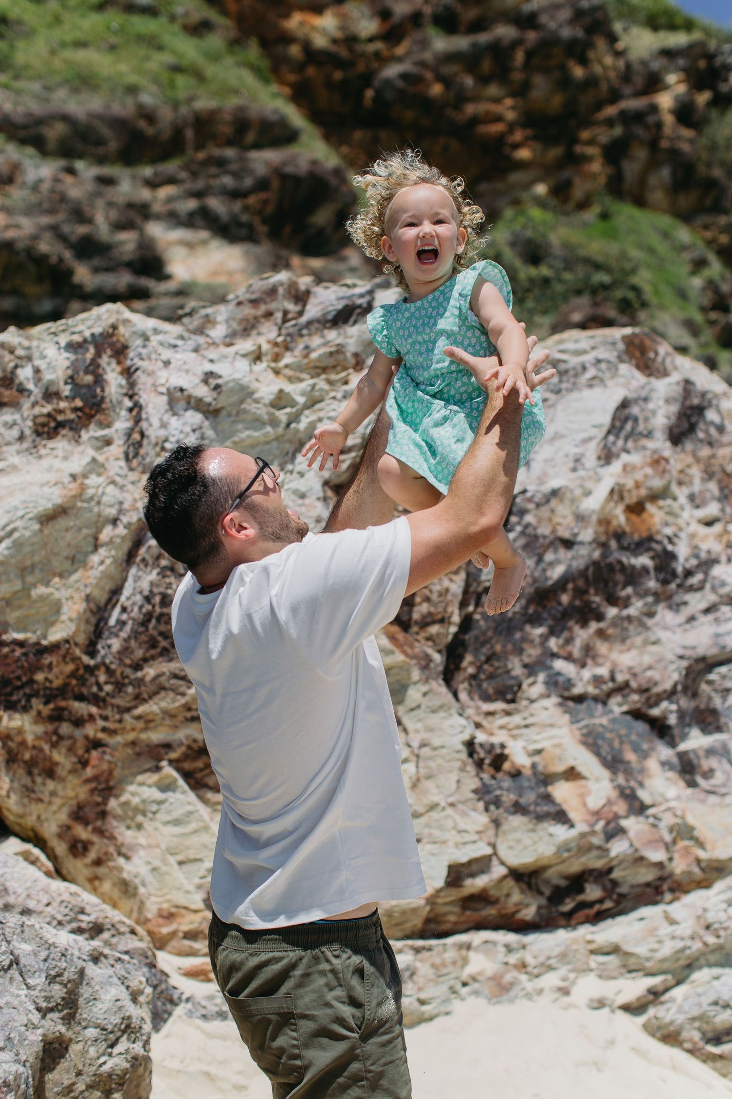 Man lifting laughing toddler girl in turquoise dress high in the air on a rocky beach.