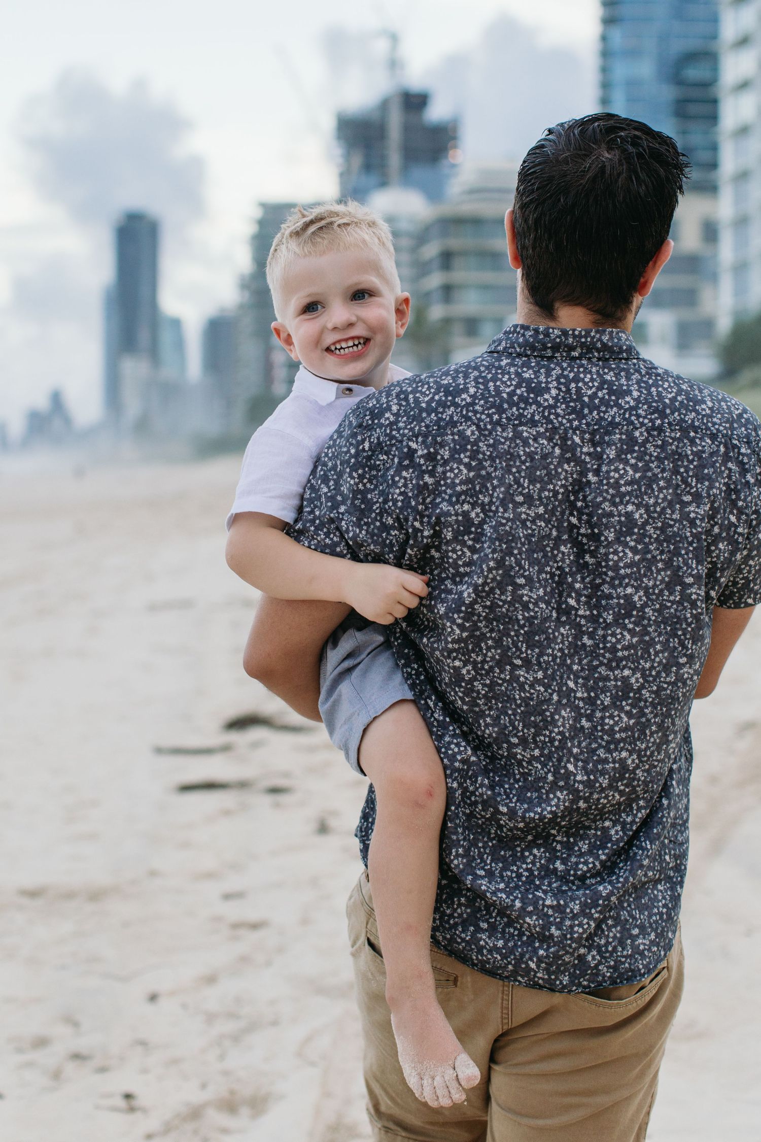 A father holds his smiling blonde toddler son on a sandy beach with city buildings in the background.