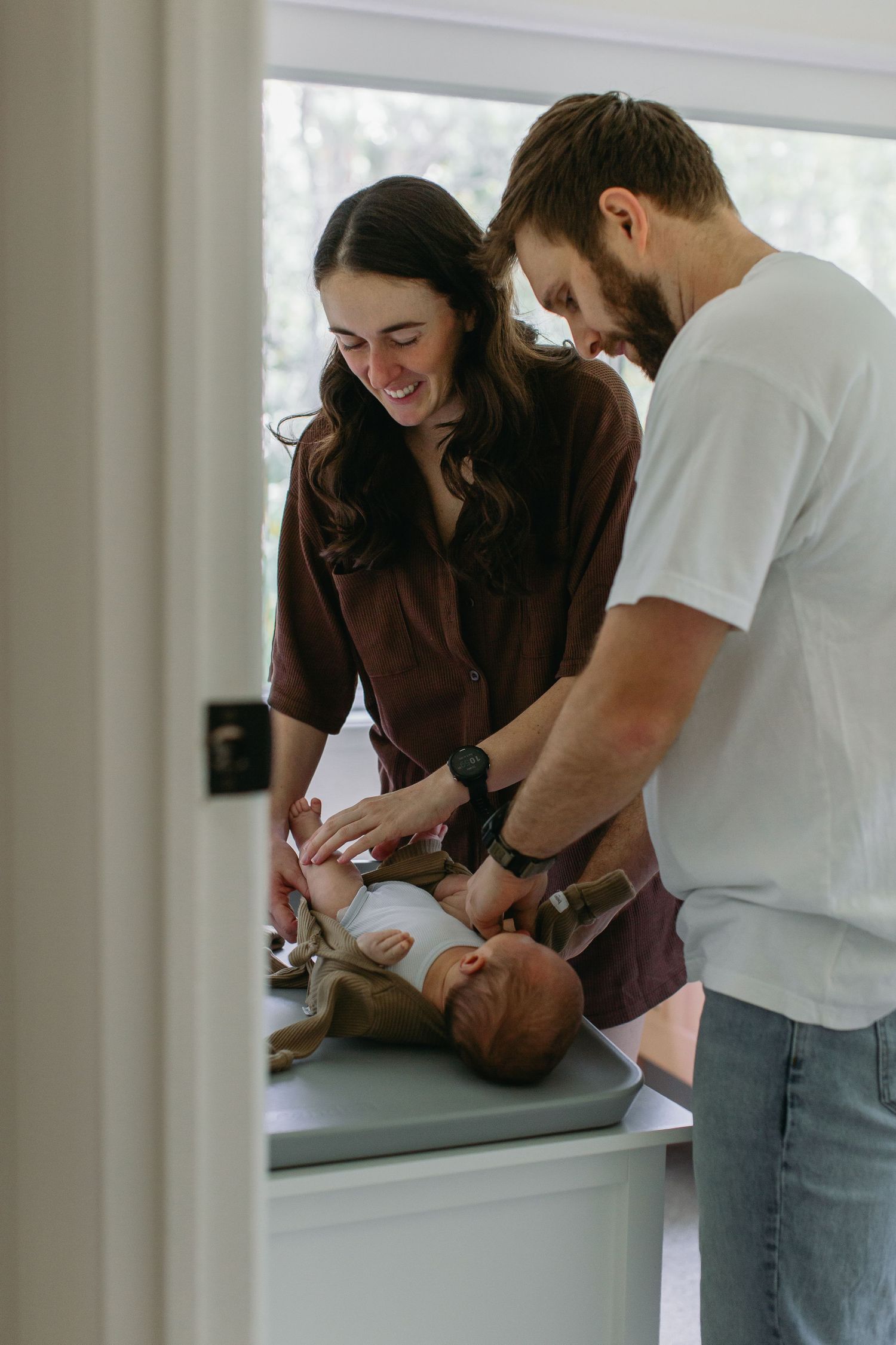 Smiling new parents lovingly changing their newborn baby on a changing table by a bright window.