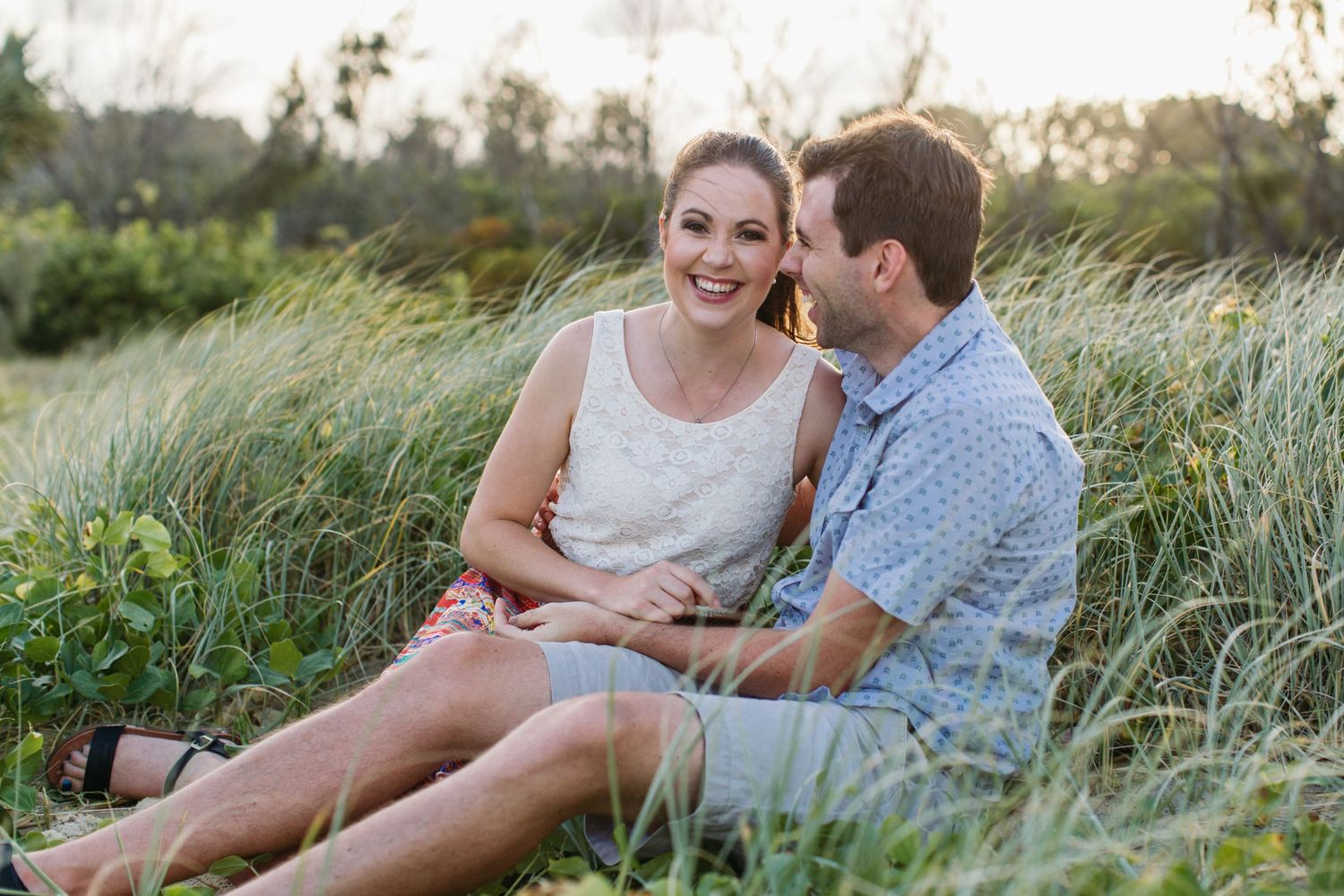 A smiling couple sits together in tall green grass during golden hour in a romantic outdoor engagement photo.