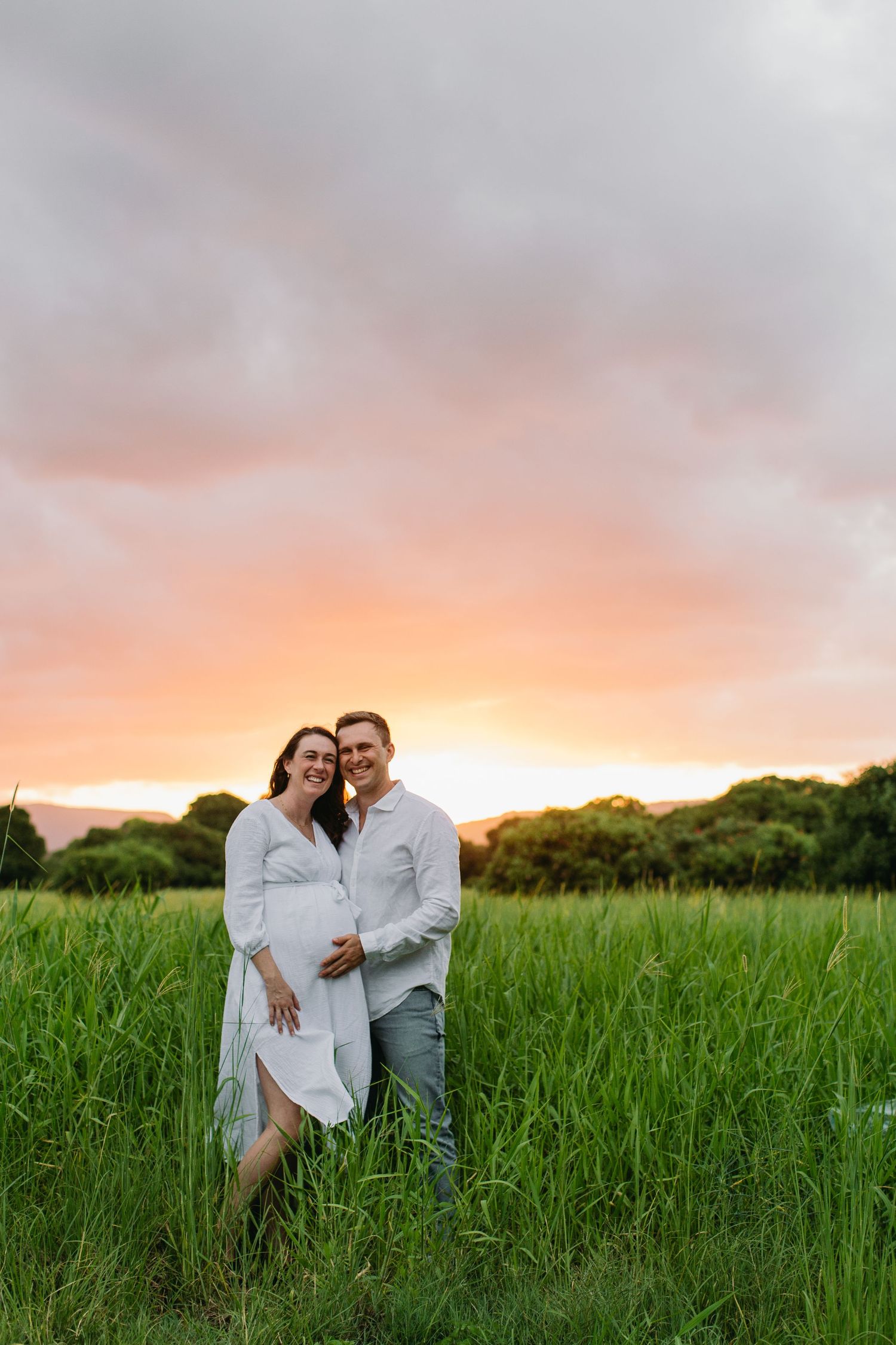 Couple posing in green field at sunset, woman in white dress with baby bump, man in white shirt, colorful sky backdrop.