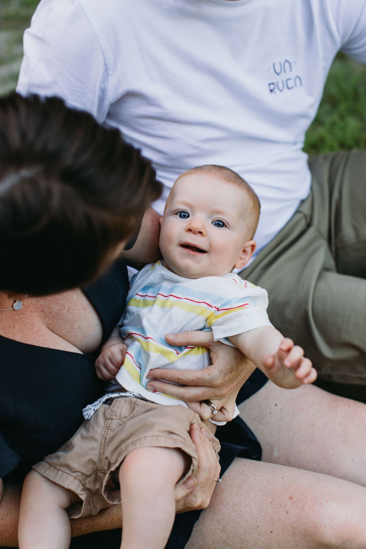 A happy baby with blue eyes sits between two adults outdoors, wearing a striped shirt and khaki shorts.