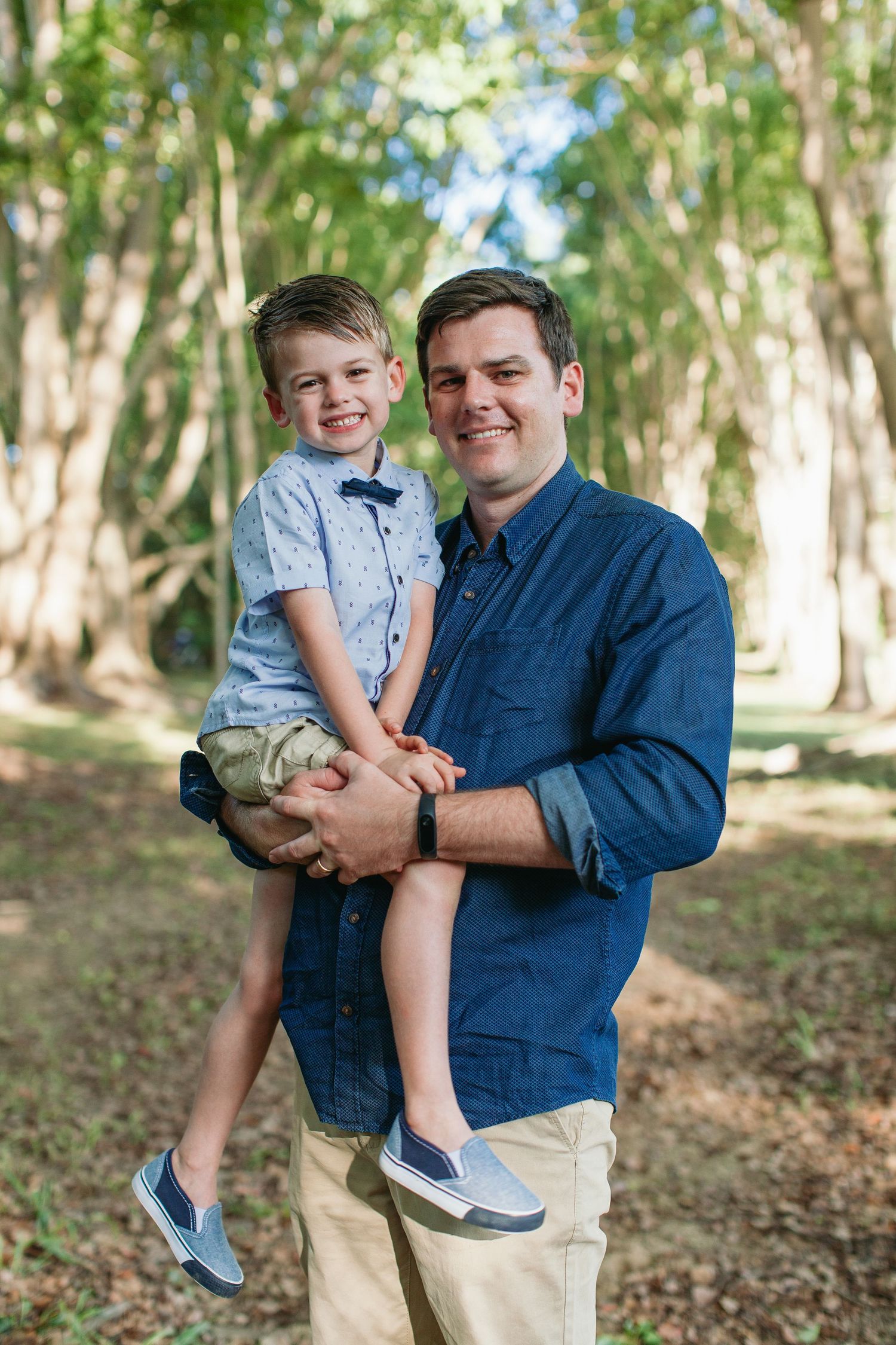 A smiling man in a blue denim shirt holds a happy young boy in a light blue shirt outdoors among tall trees.