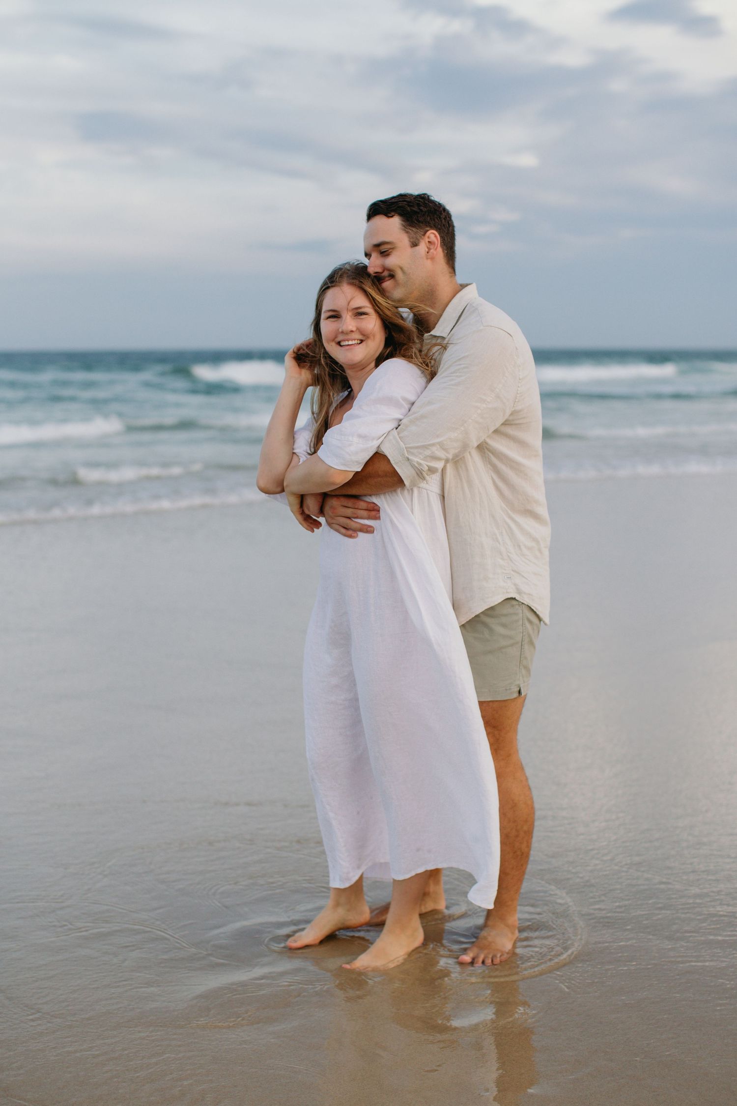 Romantic couple embracing barefoot on a wet sandy beach with gentle ocean waves behind them.