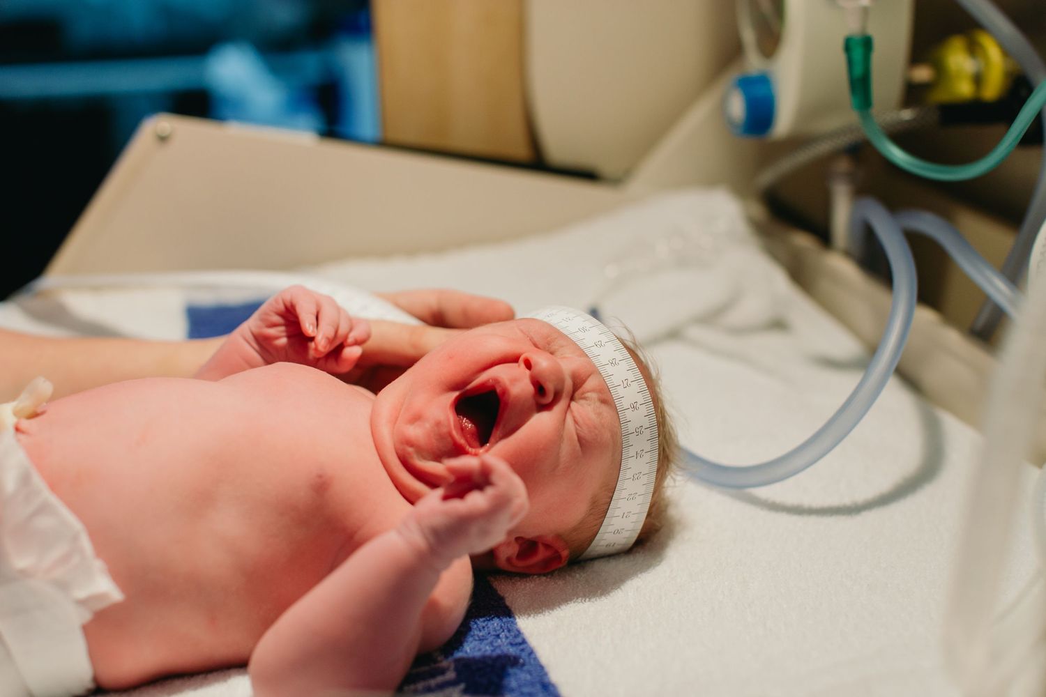 Newborn baby crying in hospital nursery with medical equipment nearby, wearing white hat after birth.