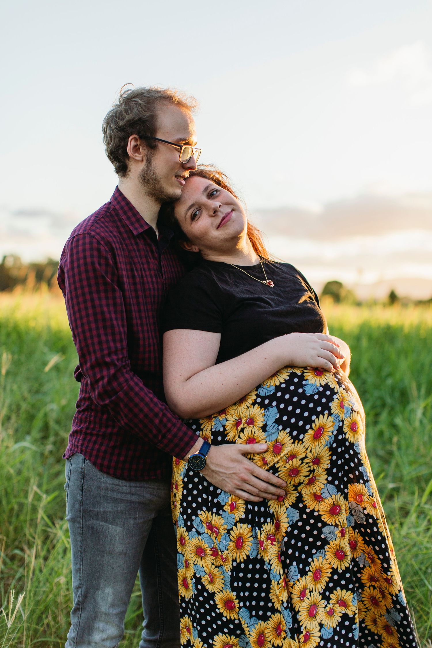 Couple embracing in a field at sunset, pregnant woman in floral skirt smiling as partner holds her bump.