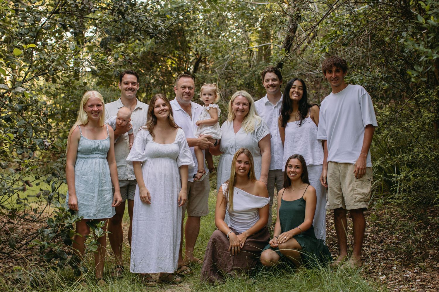 Large family group portrait smiling together in a lush green forest setting, wearing casual white and neutral outfits.