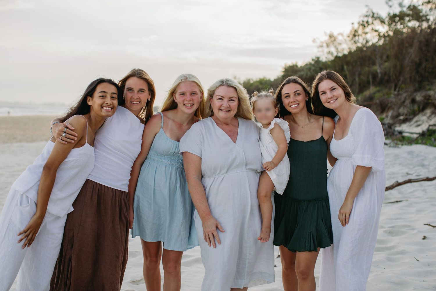 Six women smiling together on a sandy beach, wearing summer dresses in white, green, and brown tones.