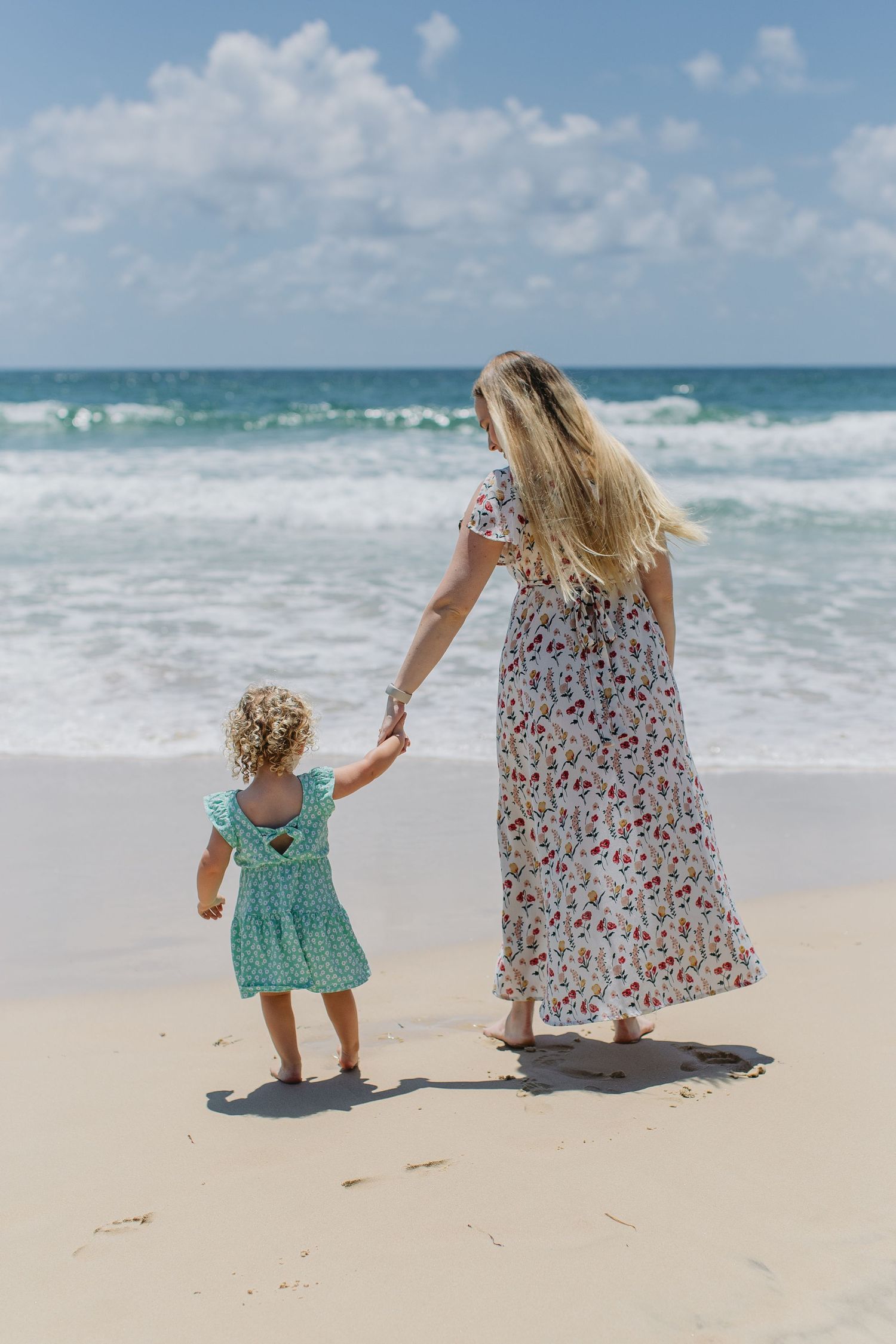 Mother and young daughter in matching floral and teal dresses walking toward ocean waves on sandy beach.