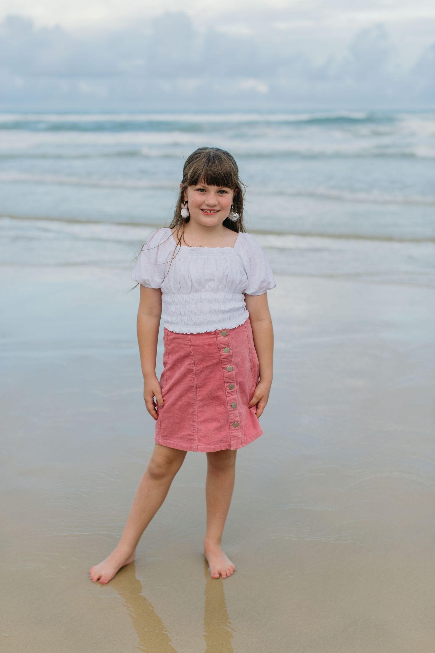 A smiling young girl in a white blouse and pink skirt poses barefoot on a sandy beach with ocean waves behind.