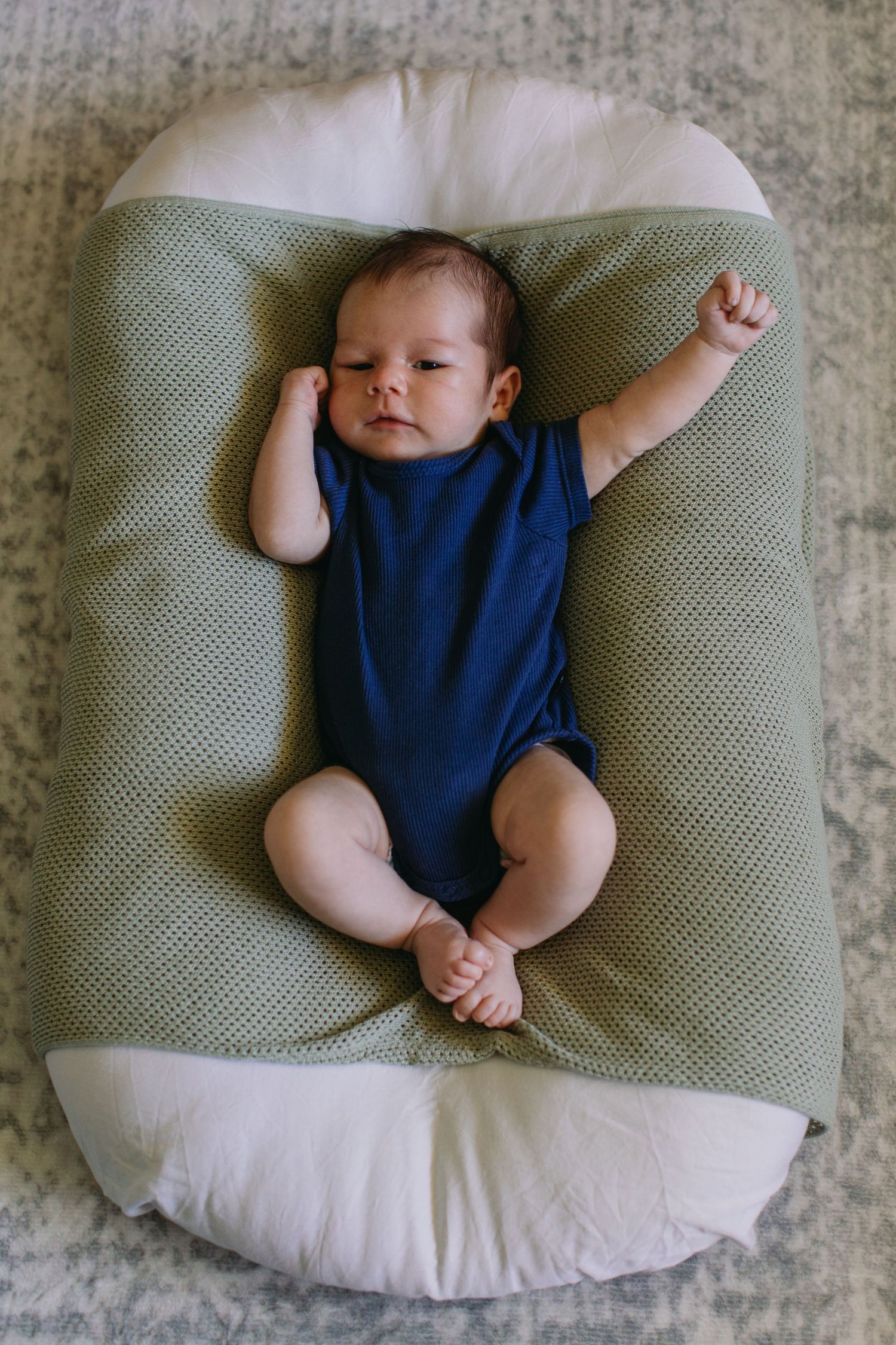 Alert newborn baby in blue onesie lying in green and white nursing pillow with one arm raised.