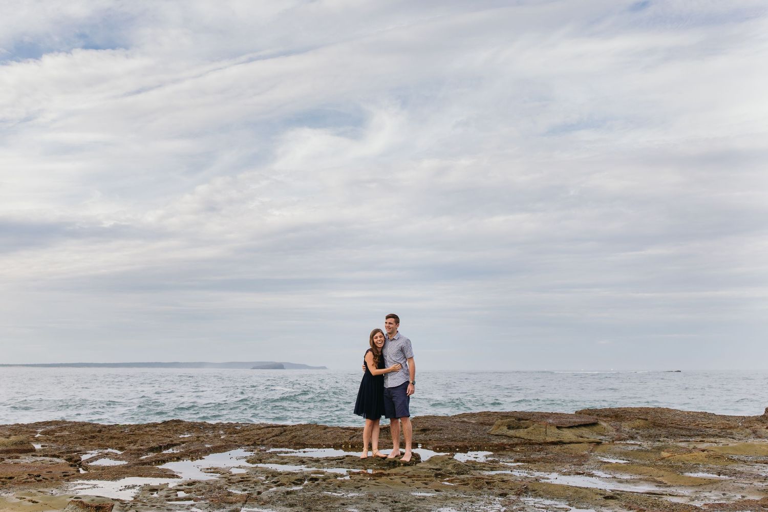 Couple embracing on rocky coastal shoreline with calm ocean and cloudy sky backdrop.
