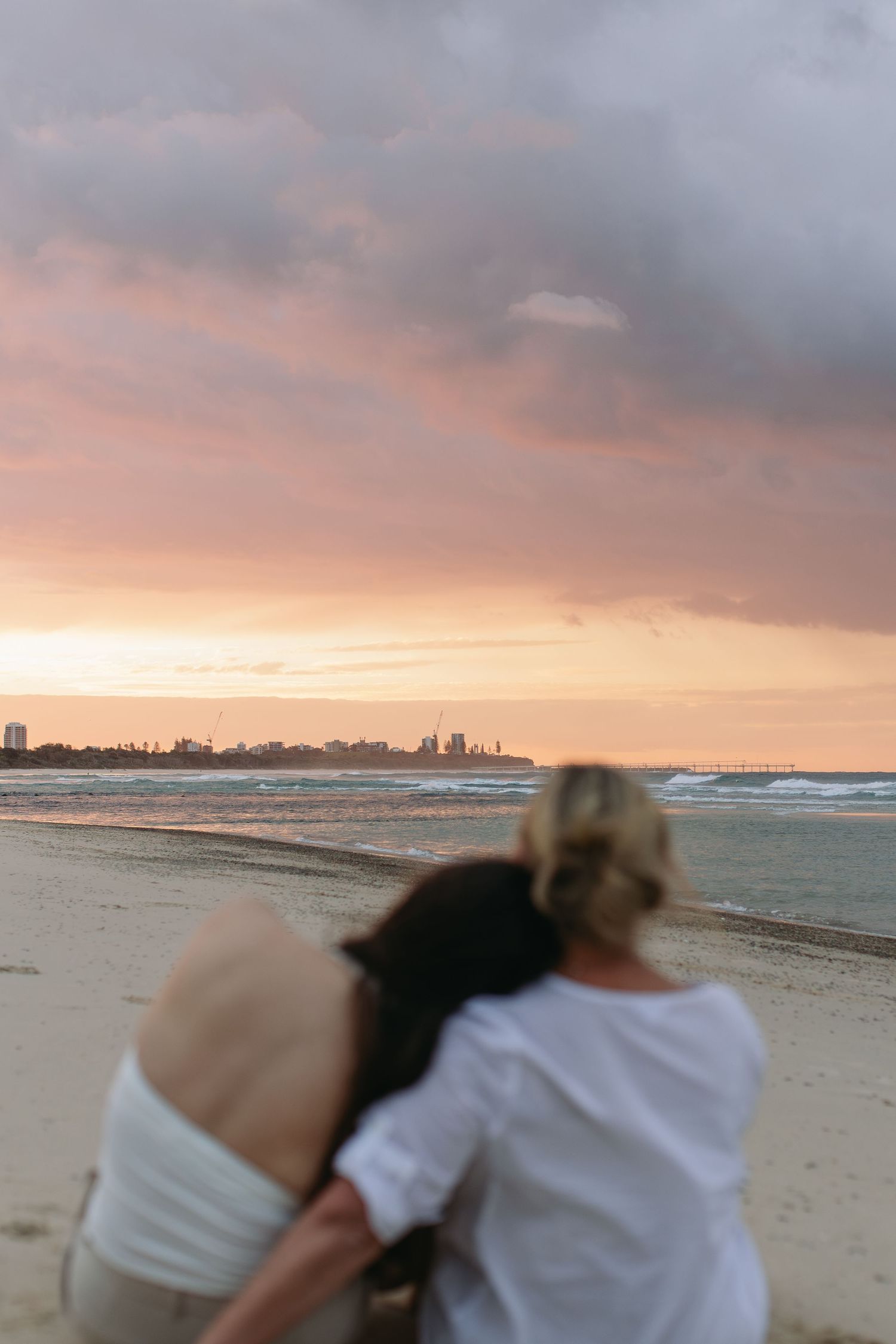 Two people embracing on a sandy beach at sunset with a city skyline and dramatic pink cloudy sky in the background.