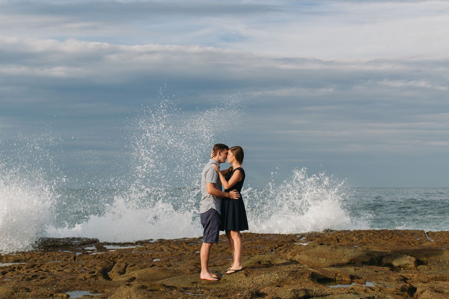 Couple kissing on rocky shore with dramatic ocean wave crashing behind them under cloudy sky.