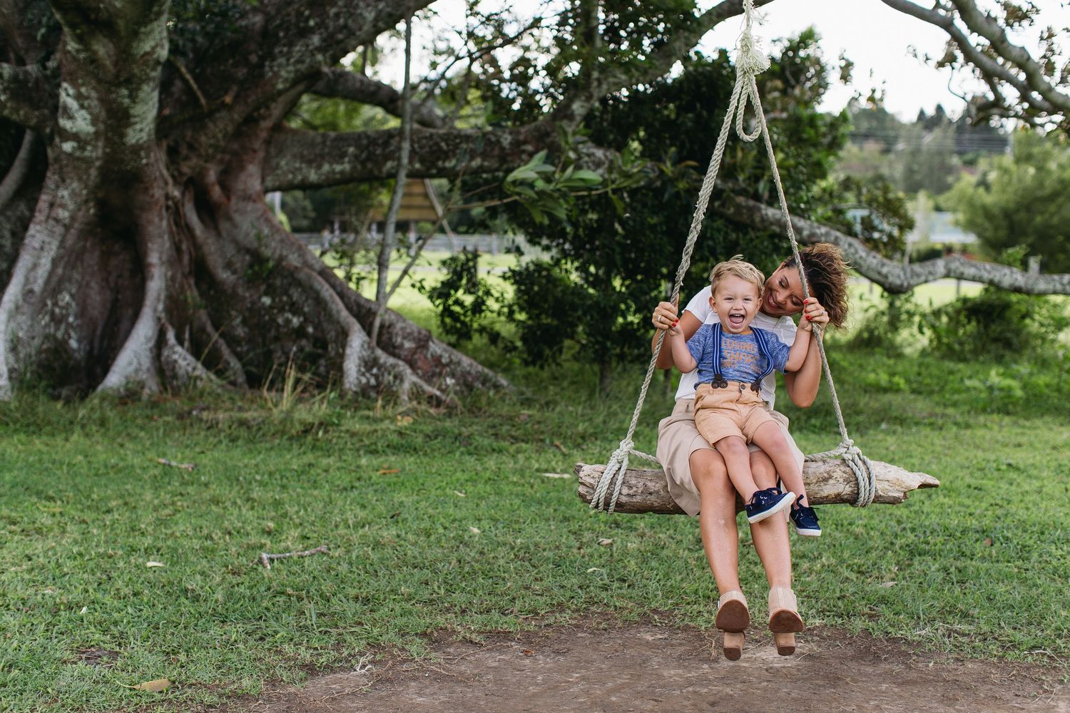 Woman pushing laughing child on rustic rope swing outdoors under large tree on green lawn.