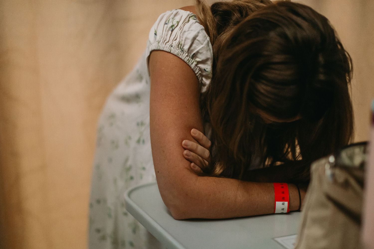 Person in hospital gown with red wristband resting head on arms at a medical facility desk.