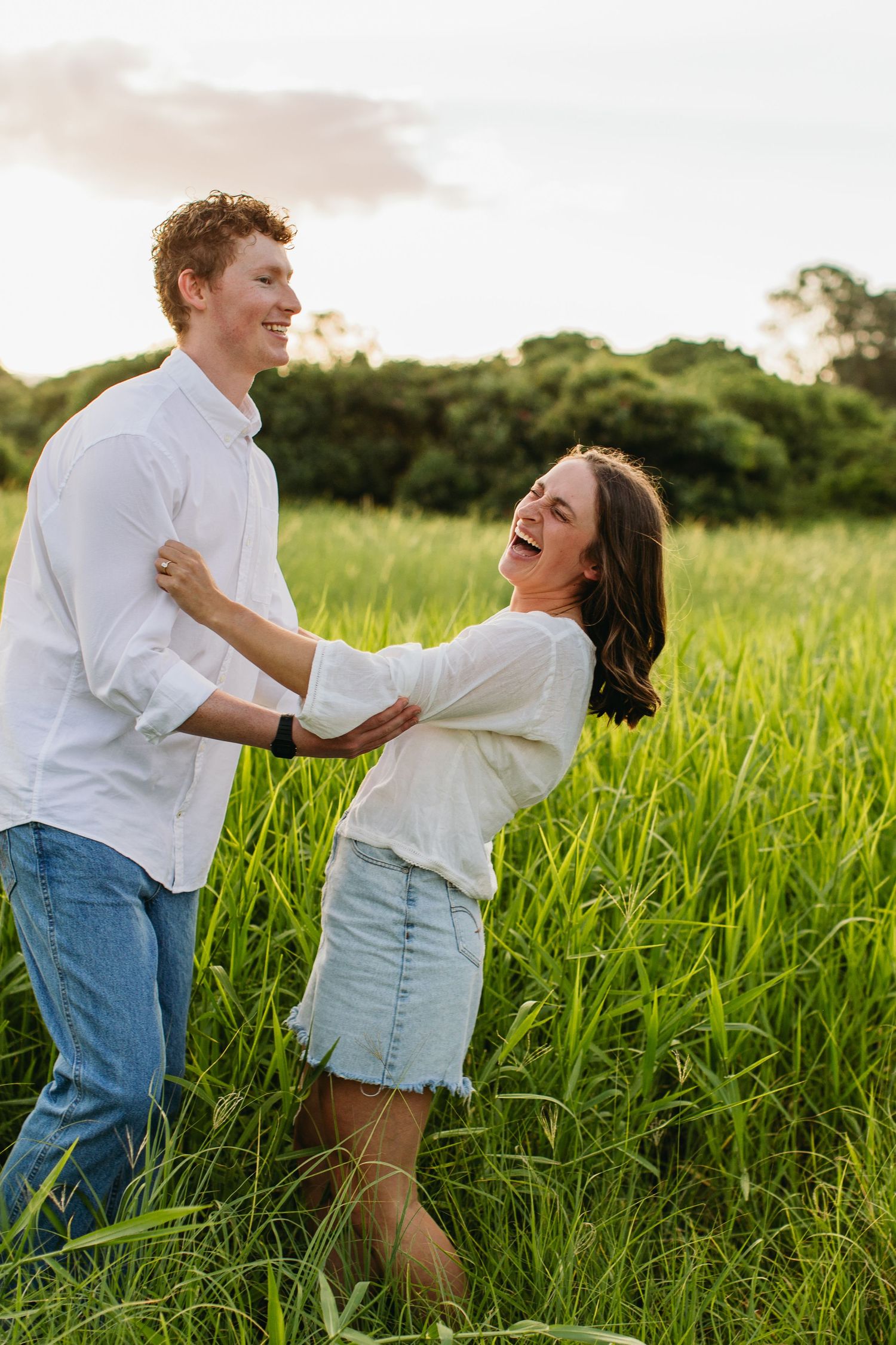 Happy couple dancing and laughing together in a lush green field at sunset, wearing white shirts and jeans.