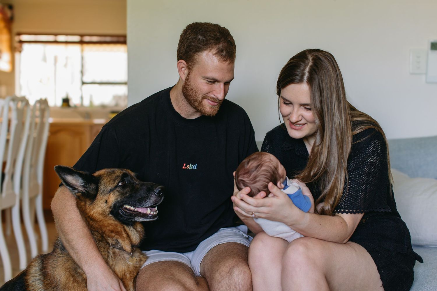 A couple sits on a couch with a German Shepherd dog, lovingly gazing at their newborn baby in a cozy home.