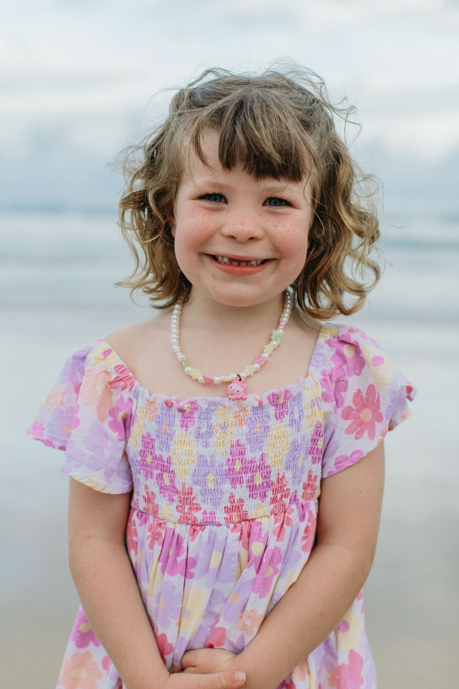 A cute young girl with curly hair in a floral dress and pearl necklace smiles on the beach.
