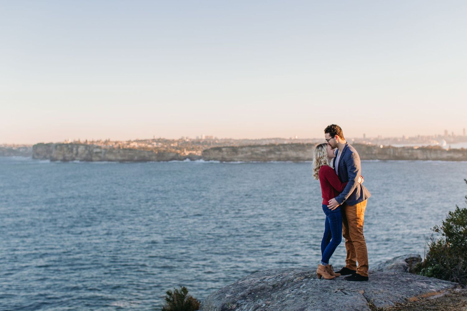Couple sharing a romantic moment on rocky cliffs overlooking a serene blue harbor at golden hour.