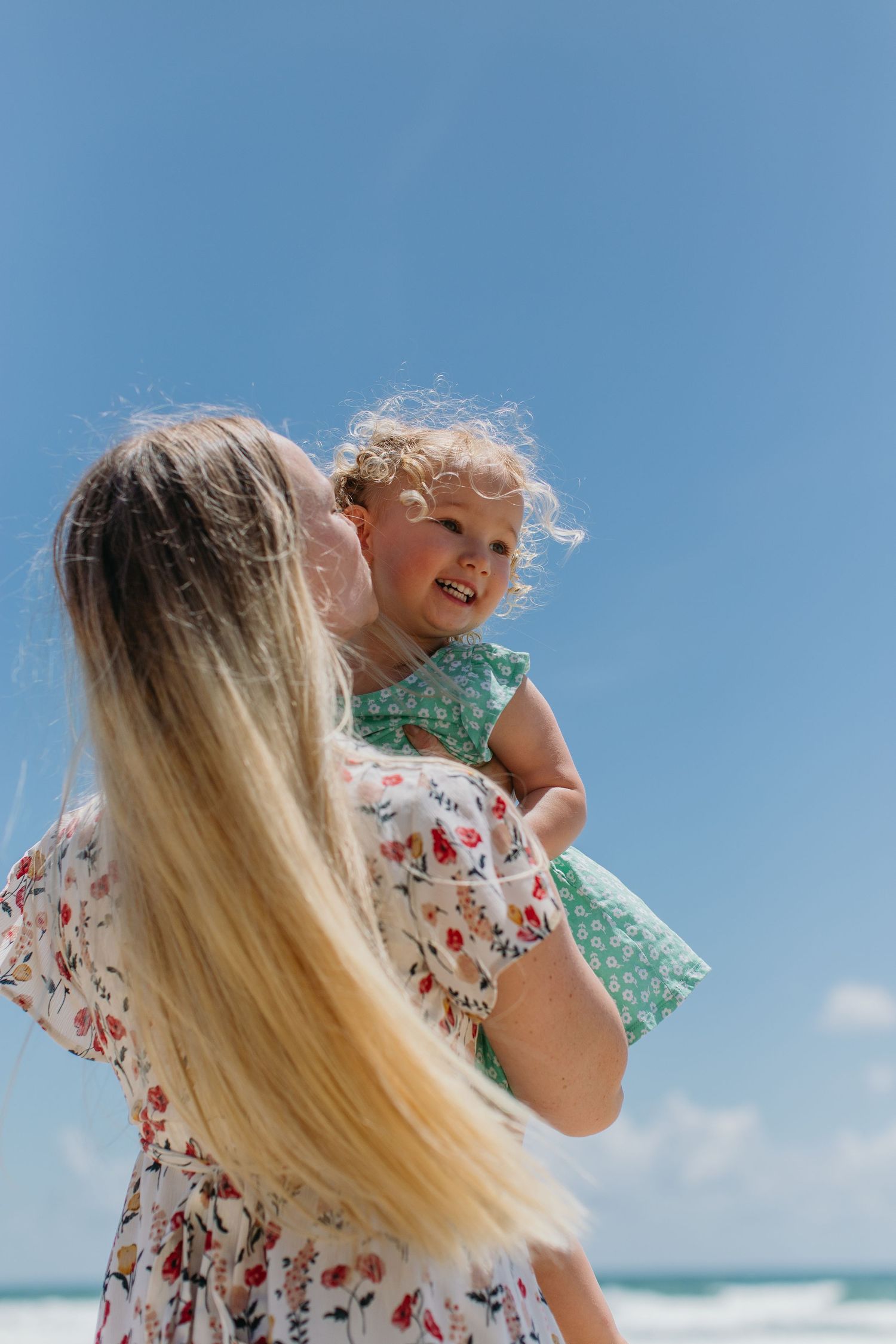 A mother holds her laughing toddler in a green dress at the beach under a bright blue sky.