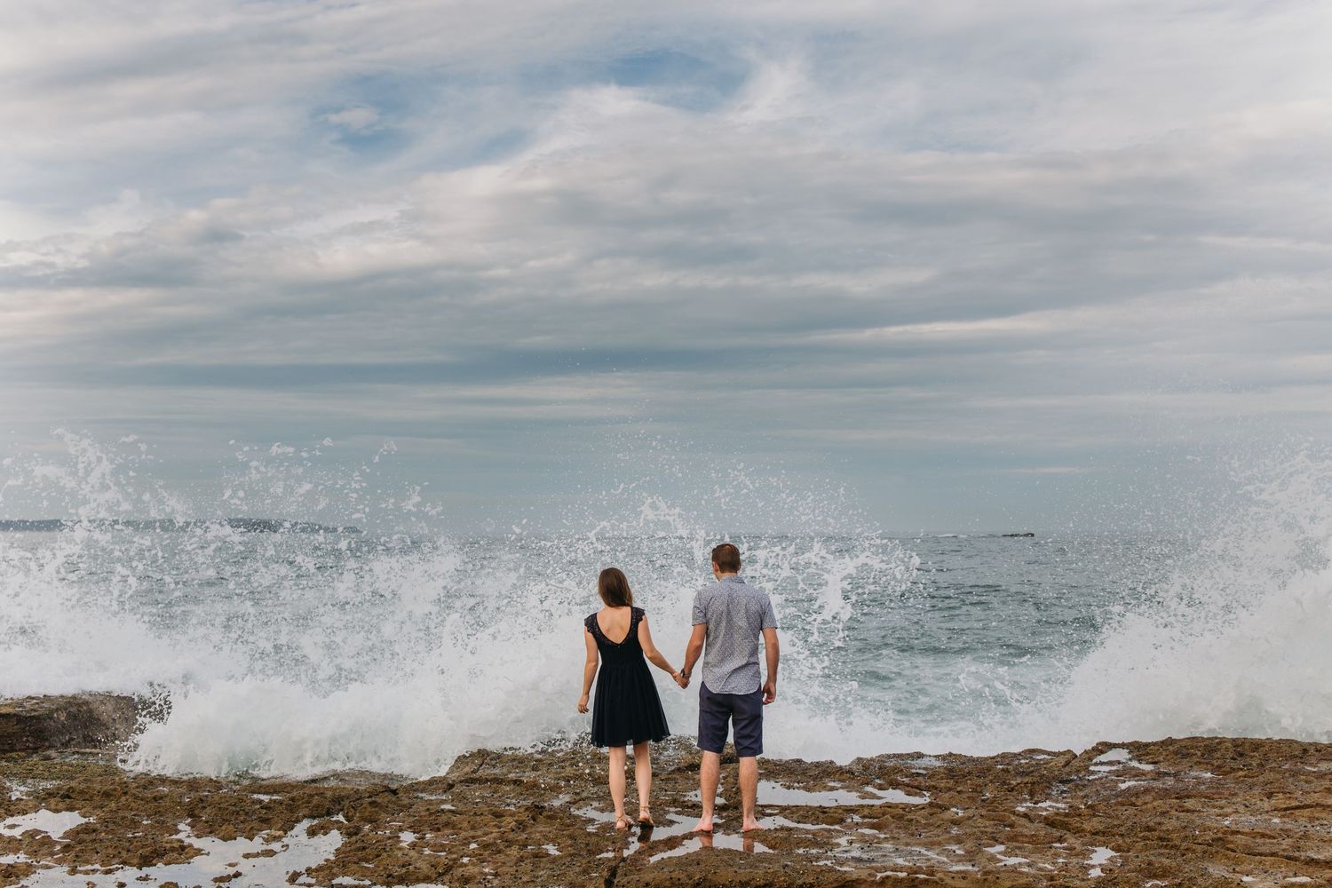 Couple holding hands on rocky shore watching dramatic ocean waves crash under cloudy sky.
