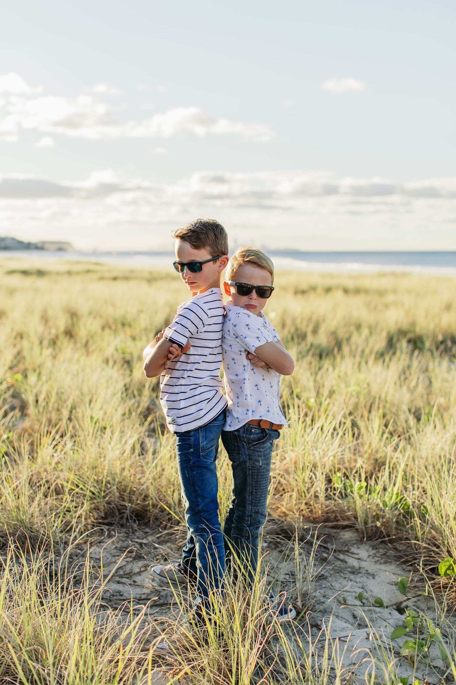 Two boys wearing sunglasses and striped shirts stand back-to-back in a coastal grass field with the ocean in the background.