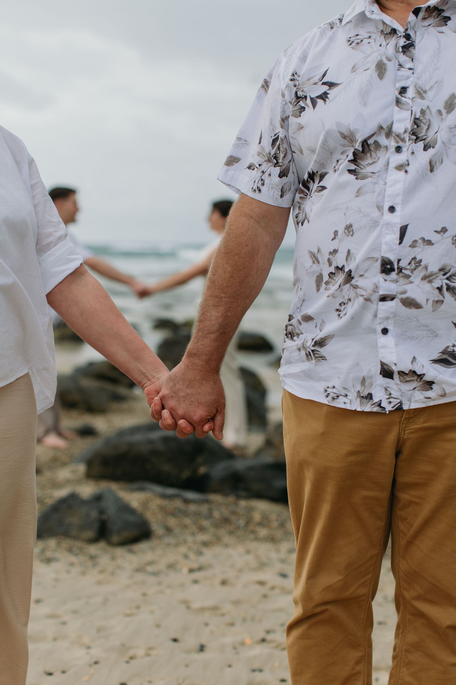 Two men holding hands on a rocky beach, wearing white shirts and tan pants, with ocean waves in the background.