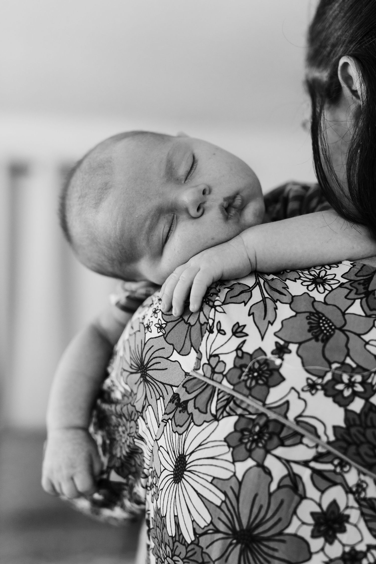 Black and white photo of a sleeping newborn baby resting on mother's shoulder wearing floral dress.