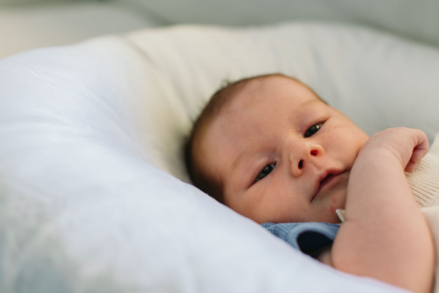 Newborn baby lying on a white pillow, looking up with curious eyes in a soft, serene setting.