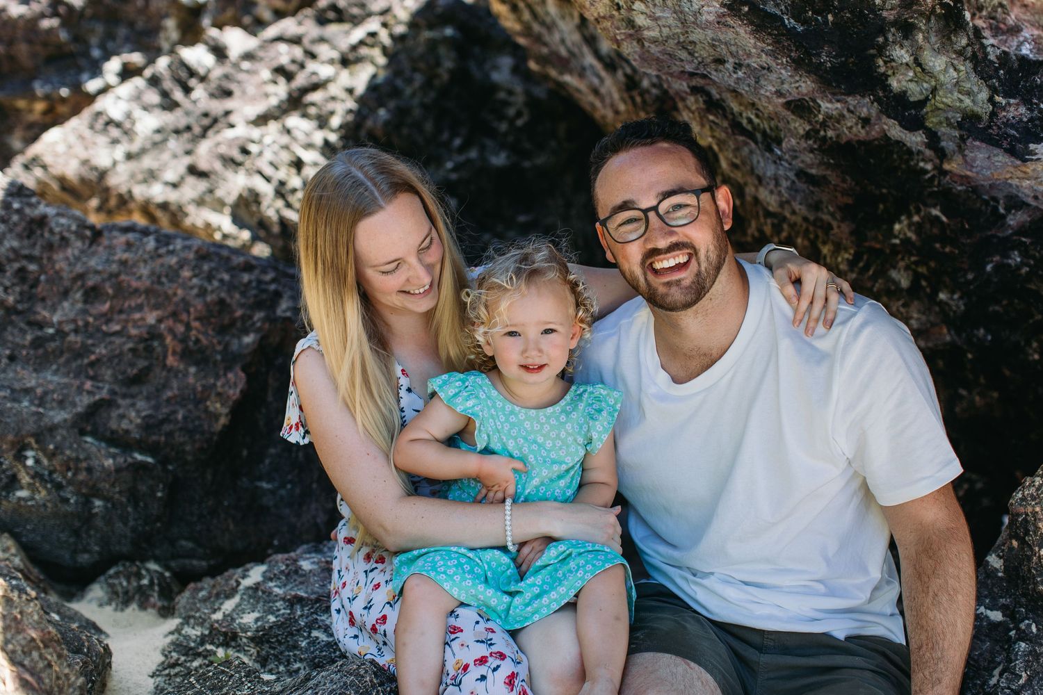 Smiling family of three sitting together on rocks at the beach on a sunny day.