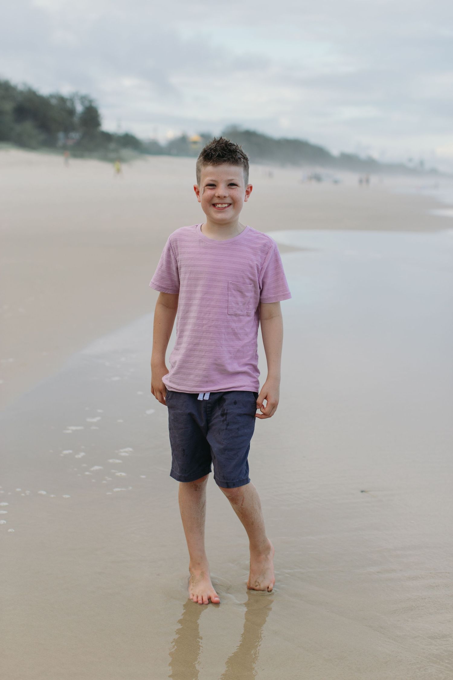 Smiling young boy in pink striped shirt and blue shorts standing barefoot on a sandy beach.