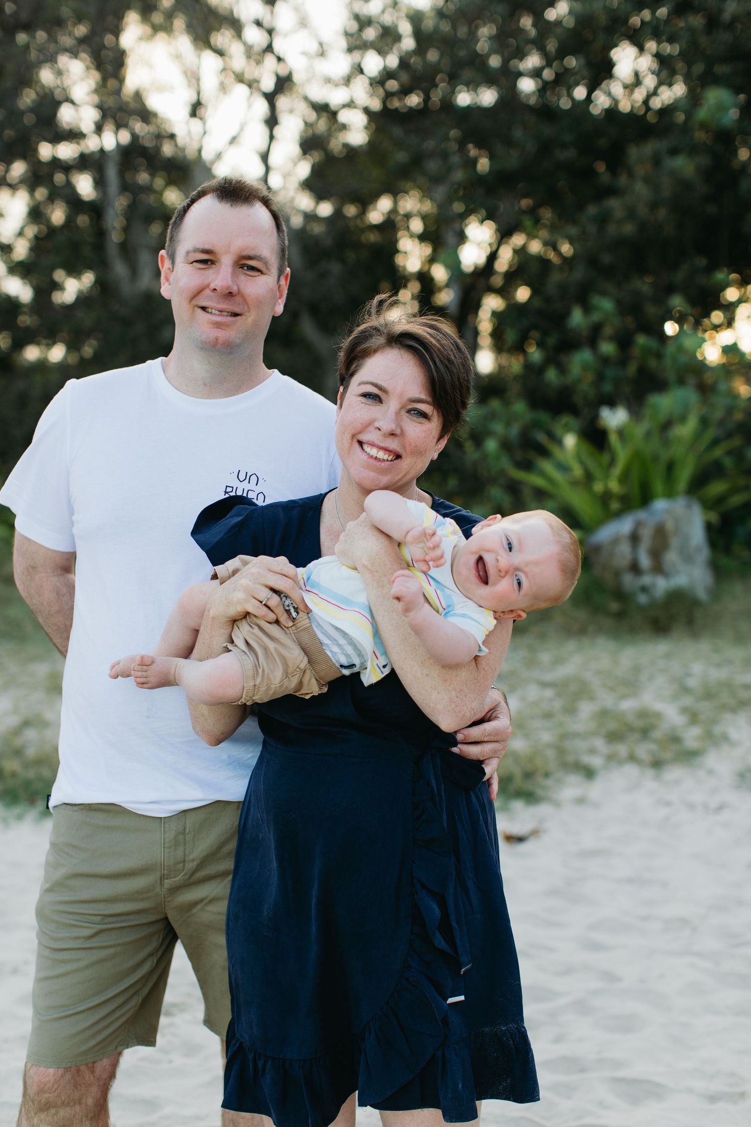 Happy family of three posing on a sandy beach, parents smiling while holding a laughing baby outdoors.