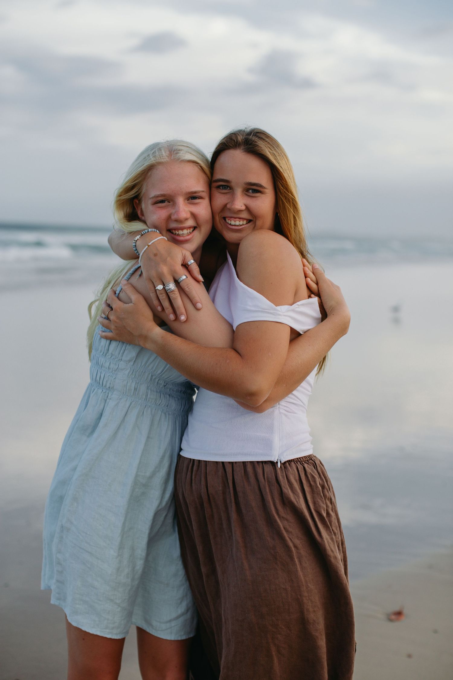 Two smiling young women hugging each other on a sandy beach with ocean waves in the background.